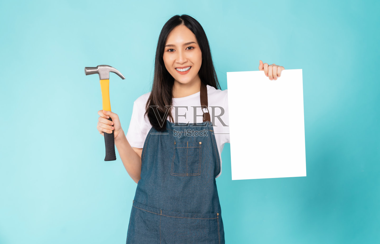 快乐的年轻亚洲妇女穿着围裙，拿着锤子，在蓝色的背景上显示一张空白的纸。照片摄影图片