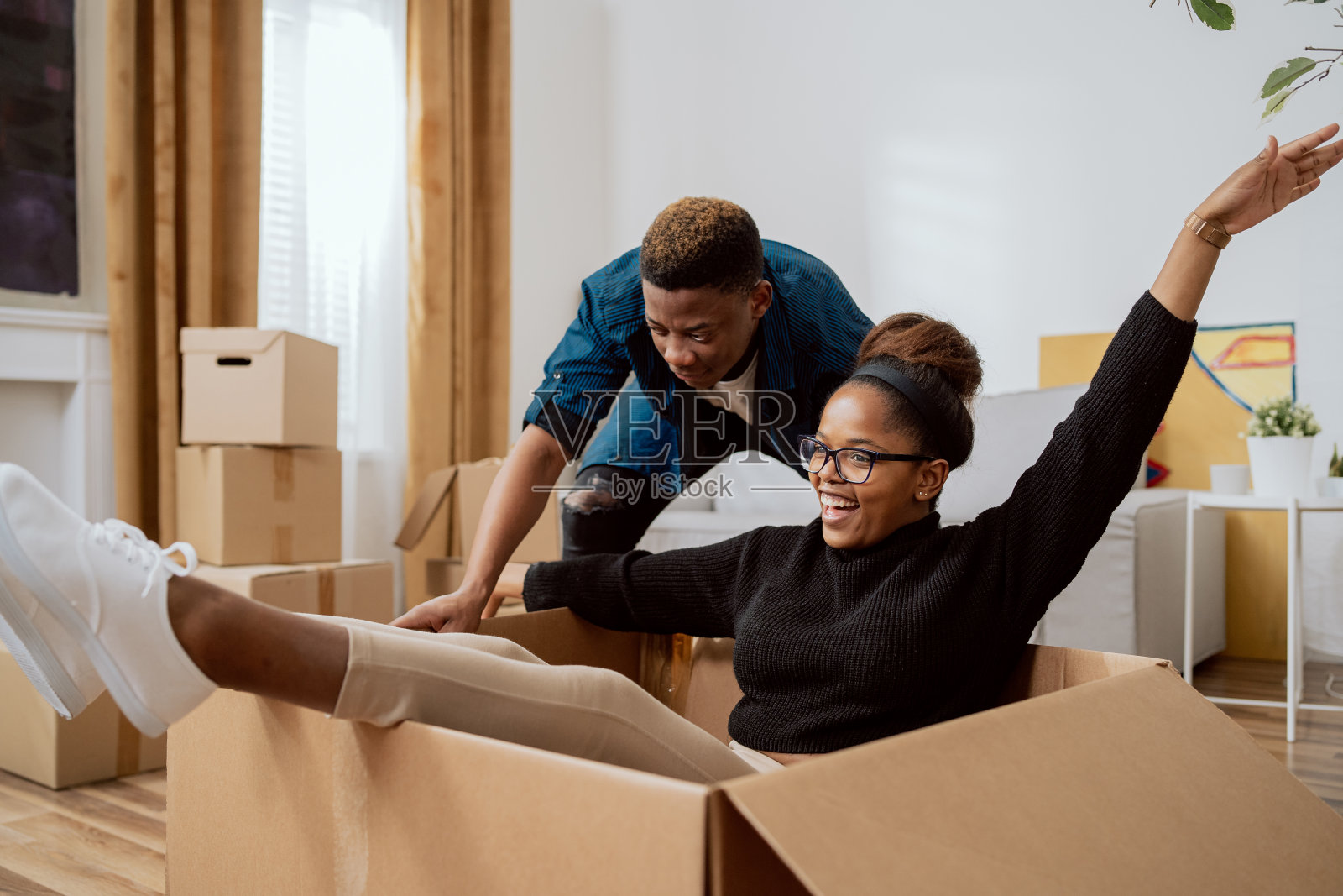 幸福的夫妇第一次买房，在搬家的日子里打开箱子玩得很开心，兴奋的妻子开车坐在纸板箱里，而丈夫推着她在新公寓里四处转转照片摄影图片
