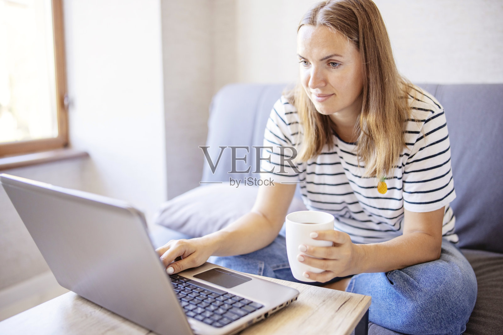 年轻迷人的女人，穿着牛仔裤和t恤，用笔记本电脑，喝着早上的咖啡。黑发女人坐在客厅里。在家工作的自由职业者照片摄影图片