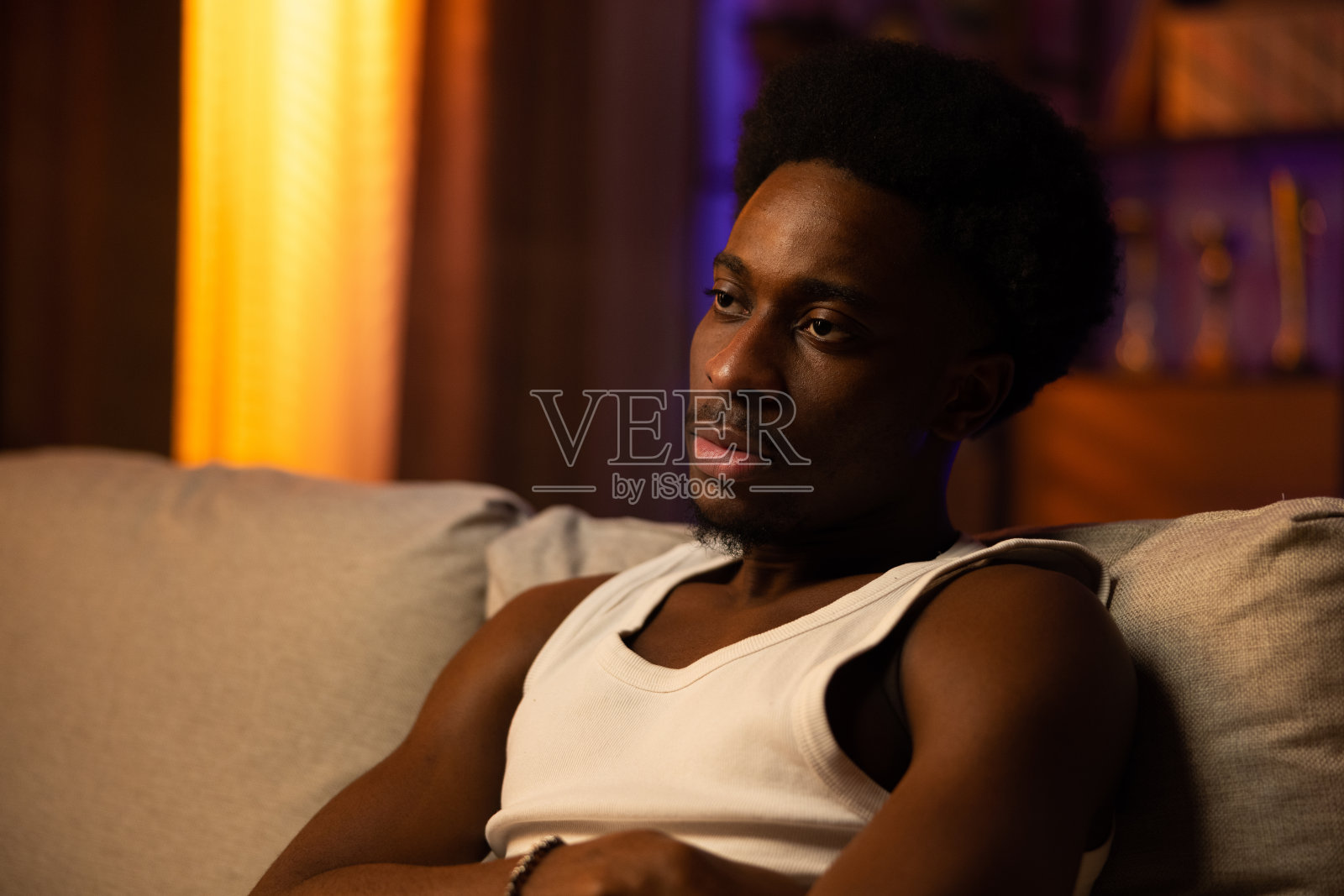 黑色皮肤的男人的肖像，胡子和爆炸头坐在沙发上，男孩穿着白色的t恤，他正在看电视，男孩的脸上有一个无聊的表情照片摄影图片