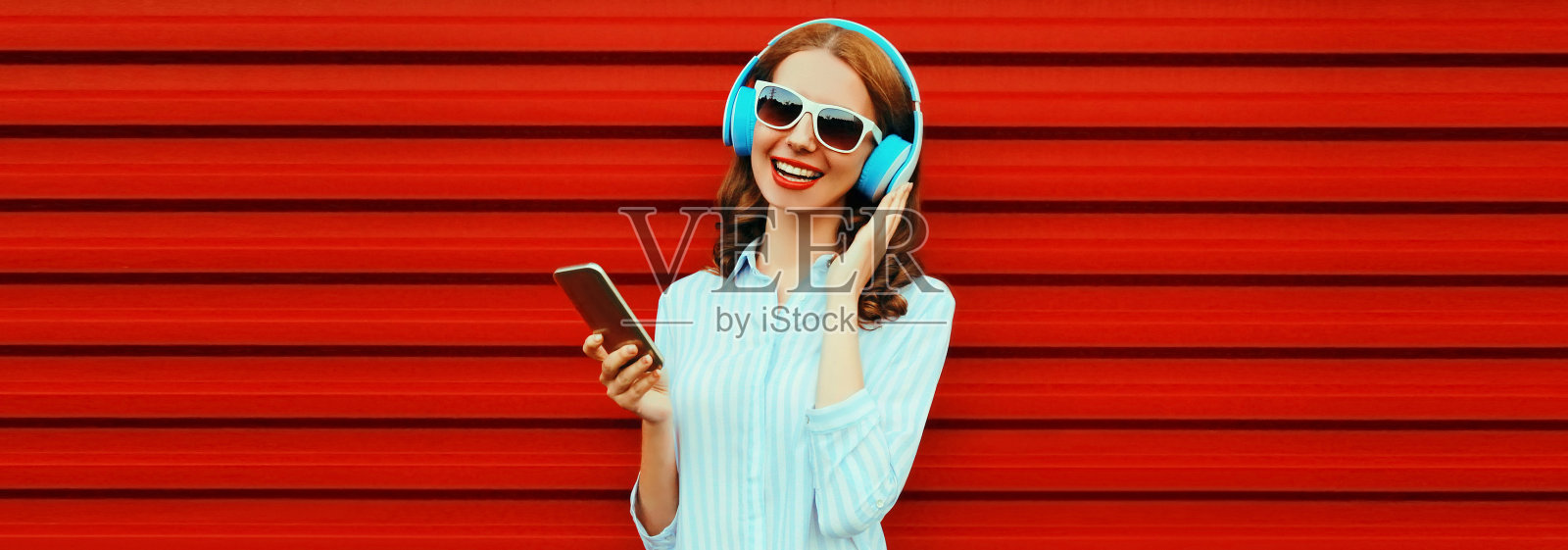 快乐微笑的年轻女子的肖像在耳机听音乐与智能手机在红色背景照片摄影图片