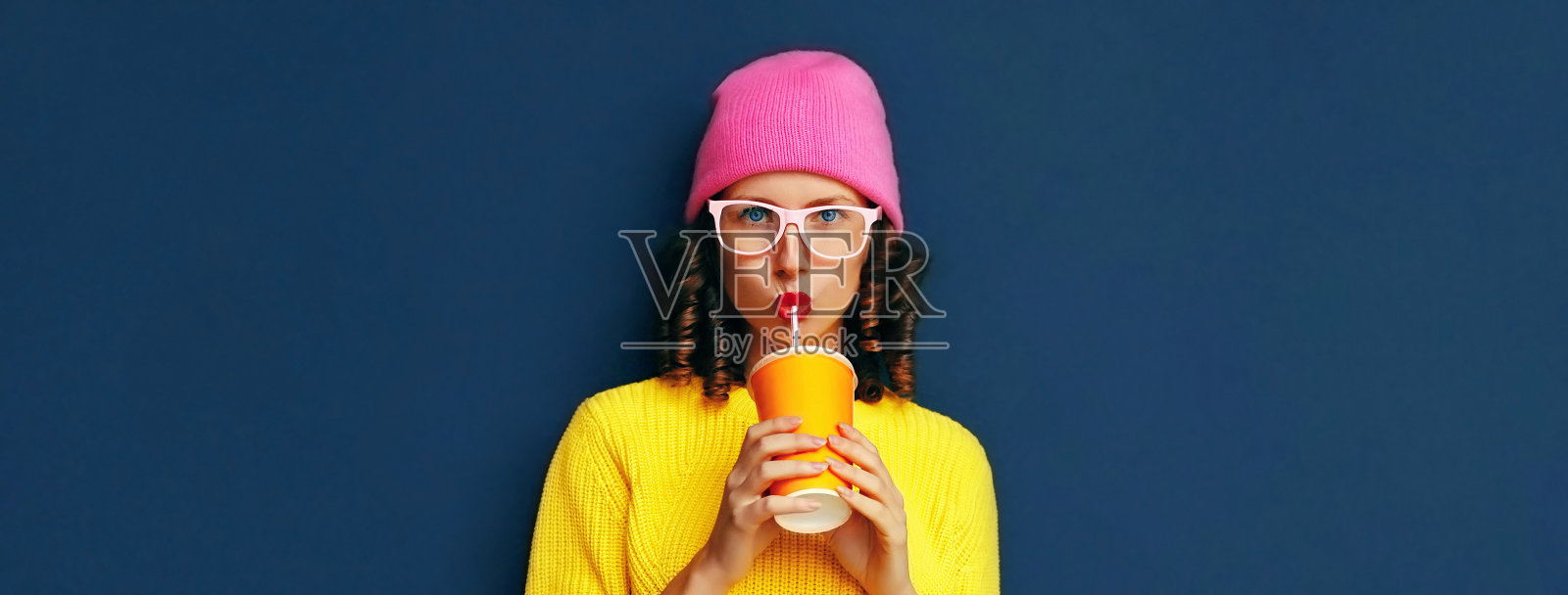 穿着鲜艳的衣服，黄色针织毛衣和粉红色帽子的时尚现代年轻女子喝鲜榨果汁的肖像，深蓝色的背景，空白的广告文字复制空间照片摄影图片