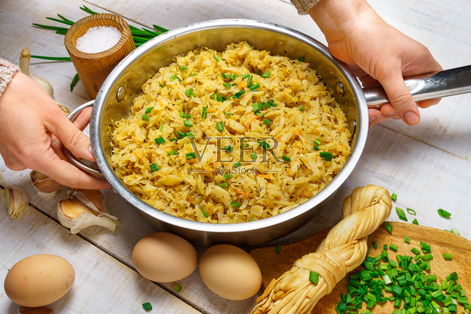 一位妇女的手在煎锅里拿着切碎的炒白菜，大蒜和大葱，奶酪，鸡蛋和盐，生的和熟的食物，在白色木板的背景上，呈现出一种乡村风格照片摄影图片