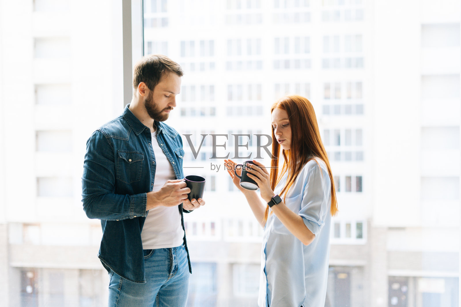 在现代的阁楼办公室里，两个年轻的同事拿着咖啡杯站在窗边讨论业务问题的肖像。照片摄影图片
