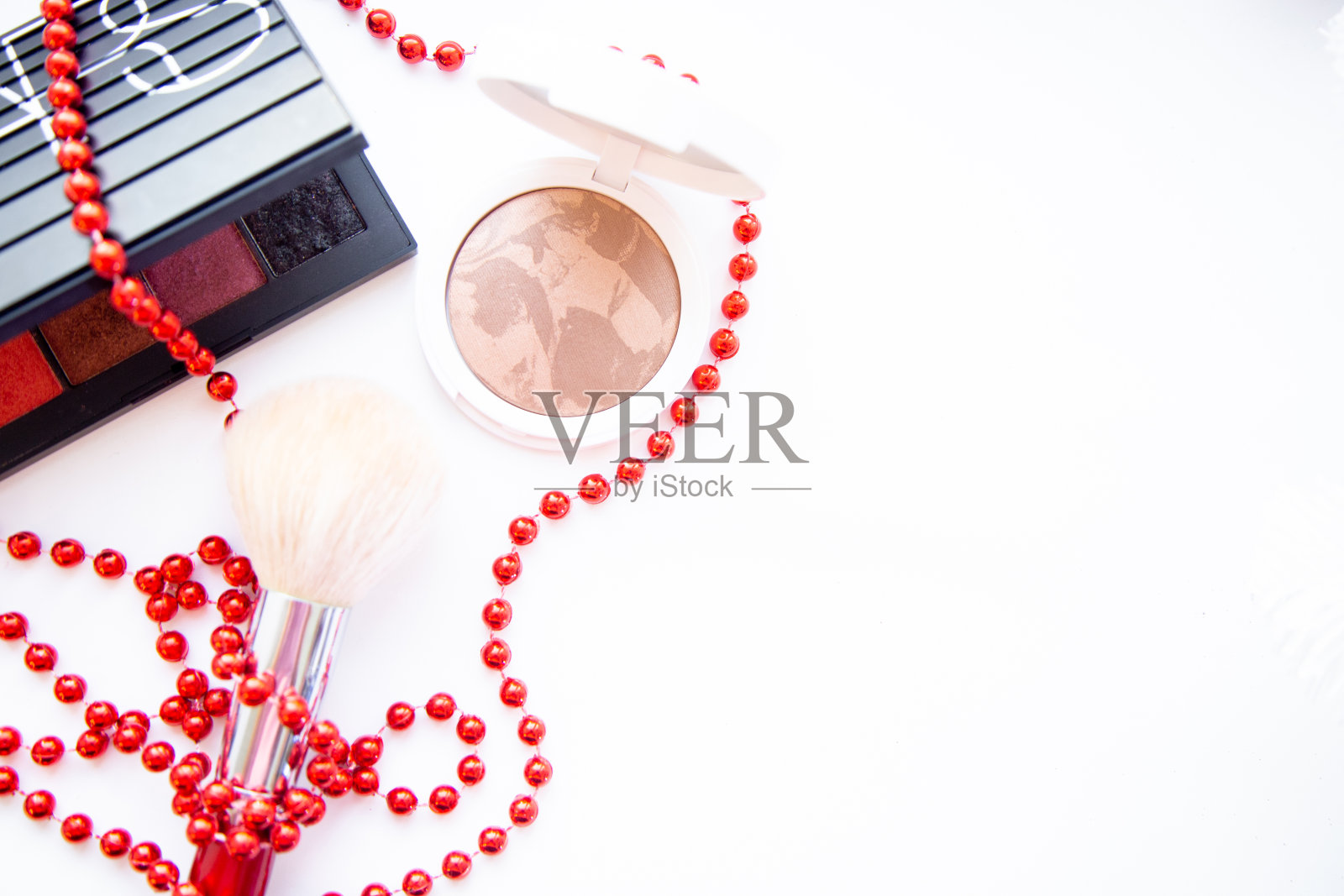 白色的粉盒和古铜色化妆品，黑色的眼影包和笔刷放在白色的背景上，它们用红色的珠子缠绕着，还有一个地方用来写文字照片摄影图片