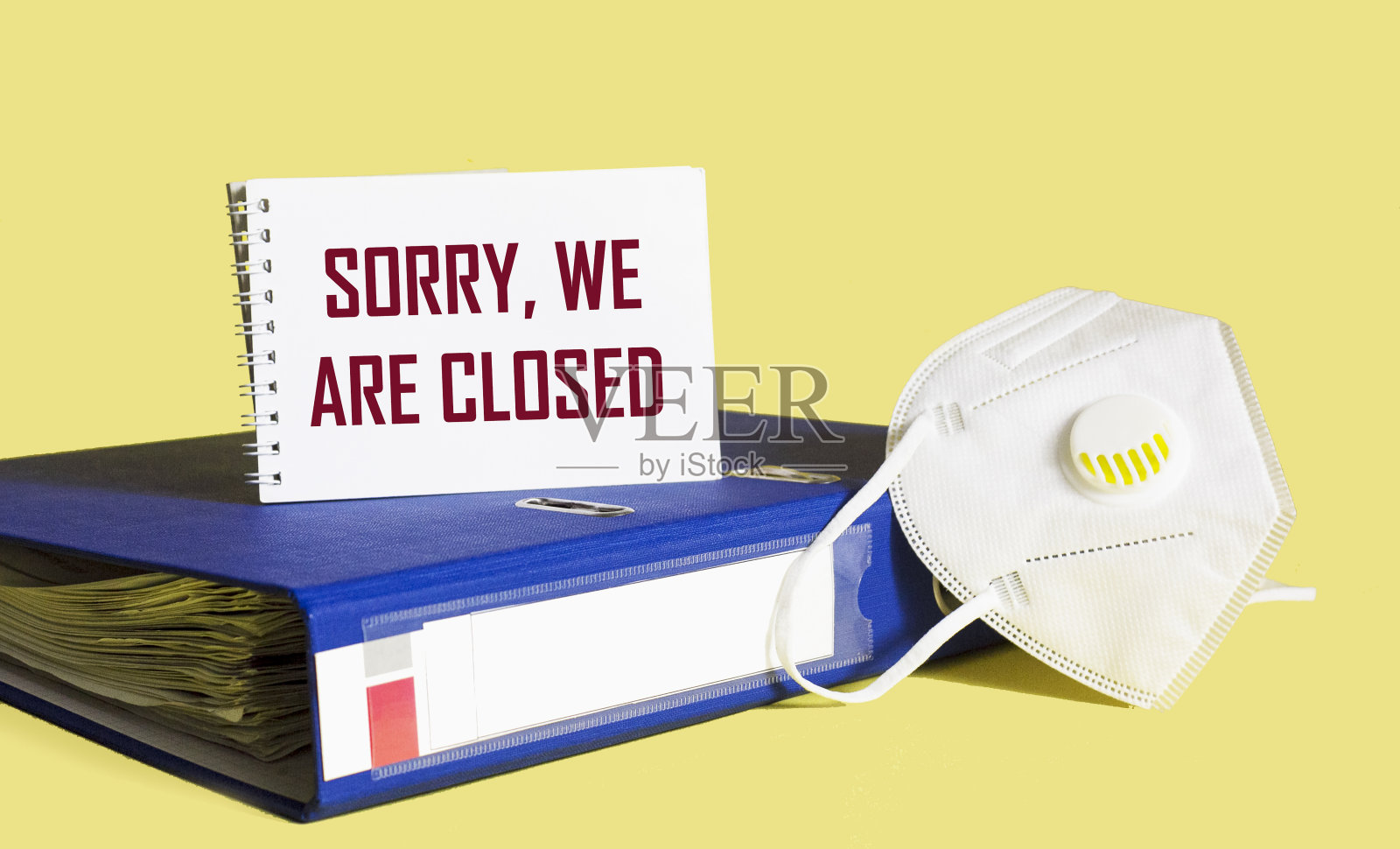 文本抱歉，我们关闭在一个笔记本和黄色背景与医疗口罩和文件夹的文件。照片摄影图片