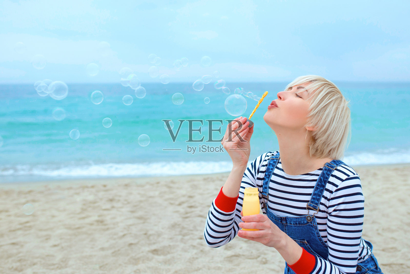 美丽的年轻漂亮的肖像，快乐，可爱，金发女人在条纹衬衫，牛仔工装裤与肥皂泡在海边照片摄影图片