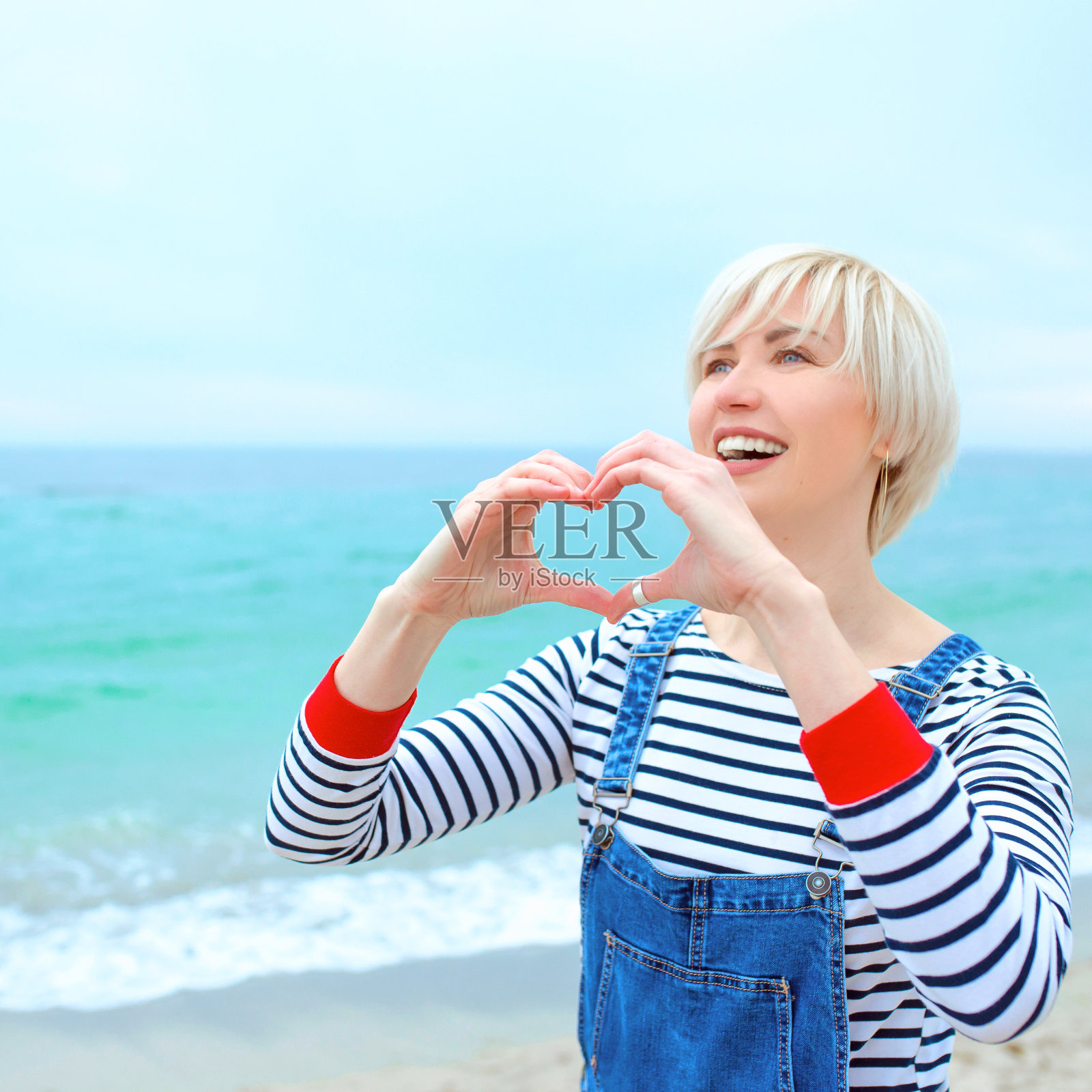 美丽的年轻金发白种人女人与手作为爱的标志度假。在令人惊叹的蓝色海洋背景下，一个女人穿着条纹衬衫，运动鞋和牛仔布工装裤站在海滩上。很高兴来到海边照片摄影图片