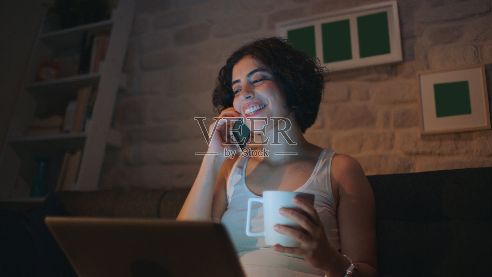 漂亮女人坐在家里用电脑工作。他一边打电话一边喝咖啡。照片摄影图片