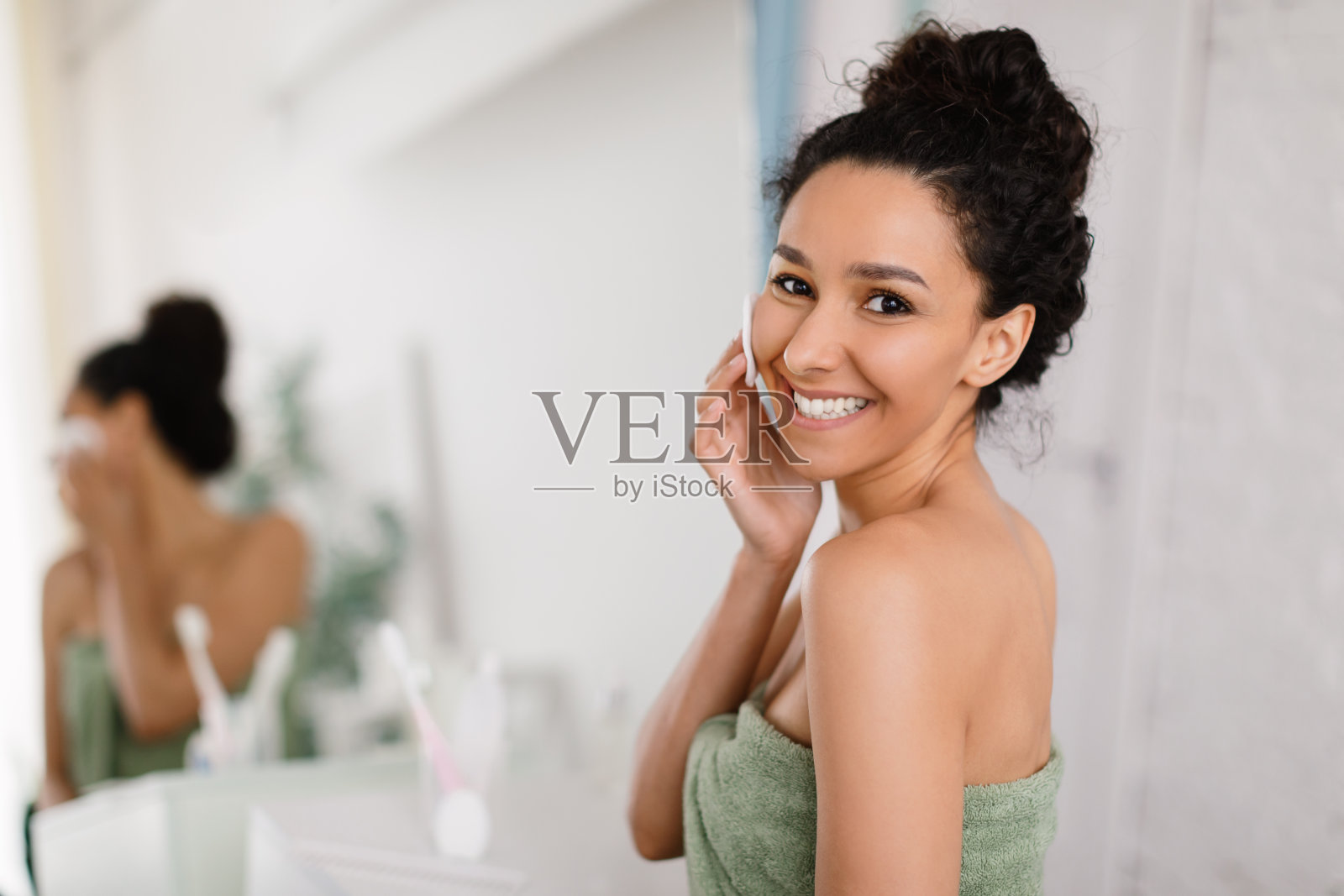 完美的皮肤。快乐的年轻黑发女人在沐浴或淋浴后用毛巾触摸脸，站在浴室的镜子旁照片摄影图片