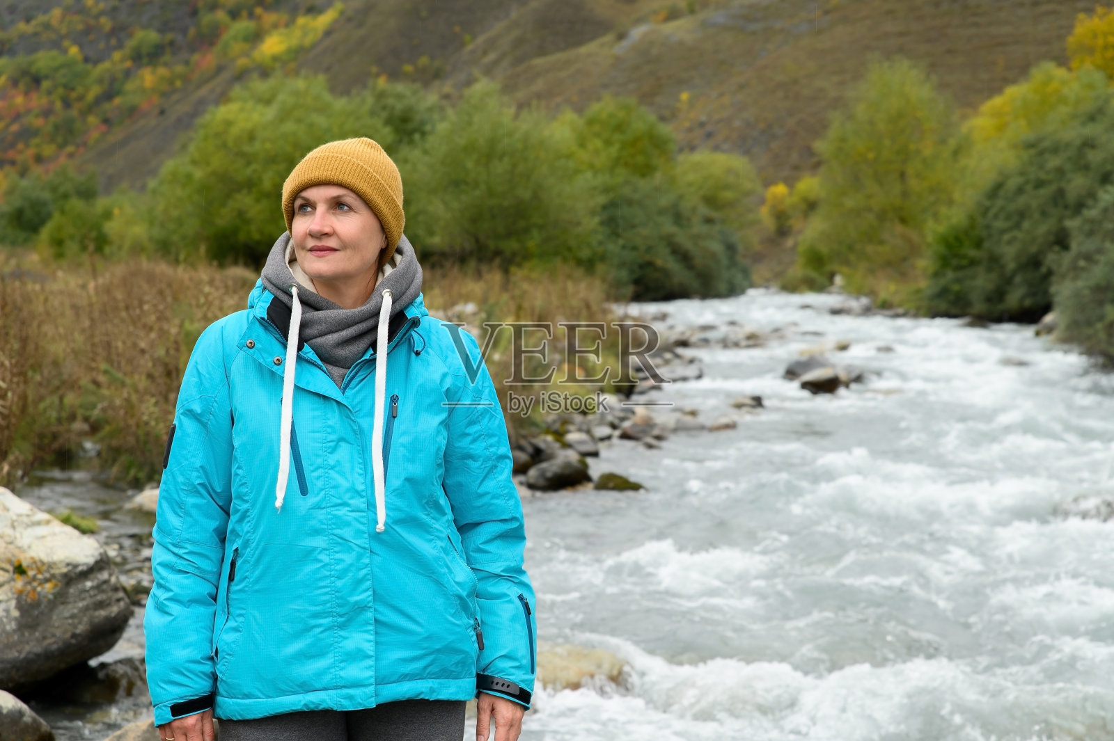 一位成年妇女穿着暖和的夹克，戴着帽子，沿着山间的河流散步照片摄影图片