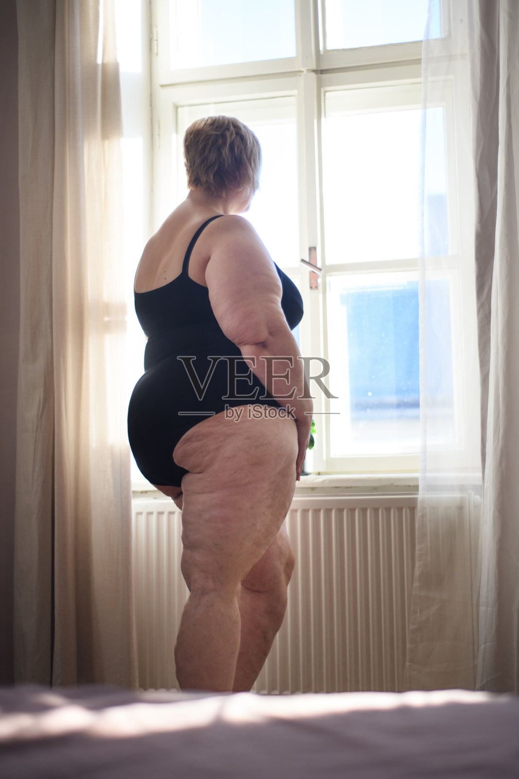 一个孤独的胖女人站在家里的窗户外看。照片摄影图片