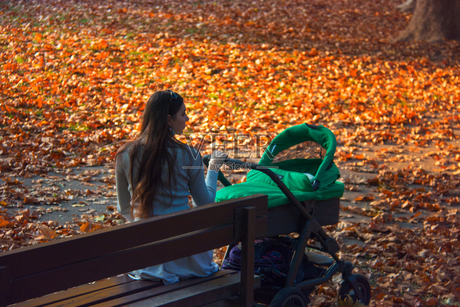 一个女人在美丽的秋日照顾一个婴儿在绿色的婴儿车，母亲坐在外面的公园木凳与彩色的秋叶背景照片摄影图片
