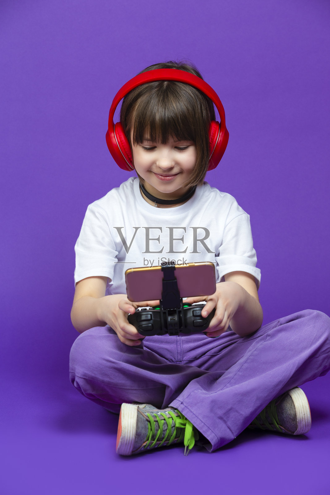 穿着白色t恤的白人小女孩用游戏手柄和红色耳机摆造型，表情惊讶地用手机玩游戏照片摄影图片