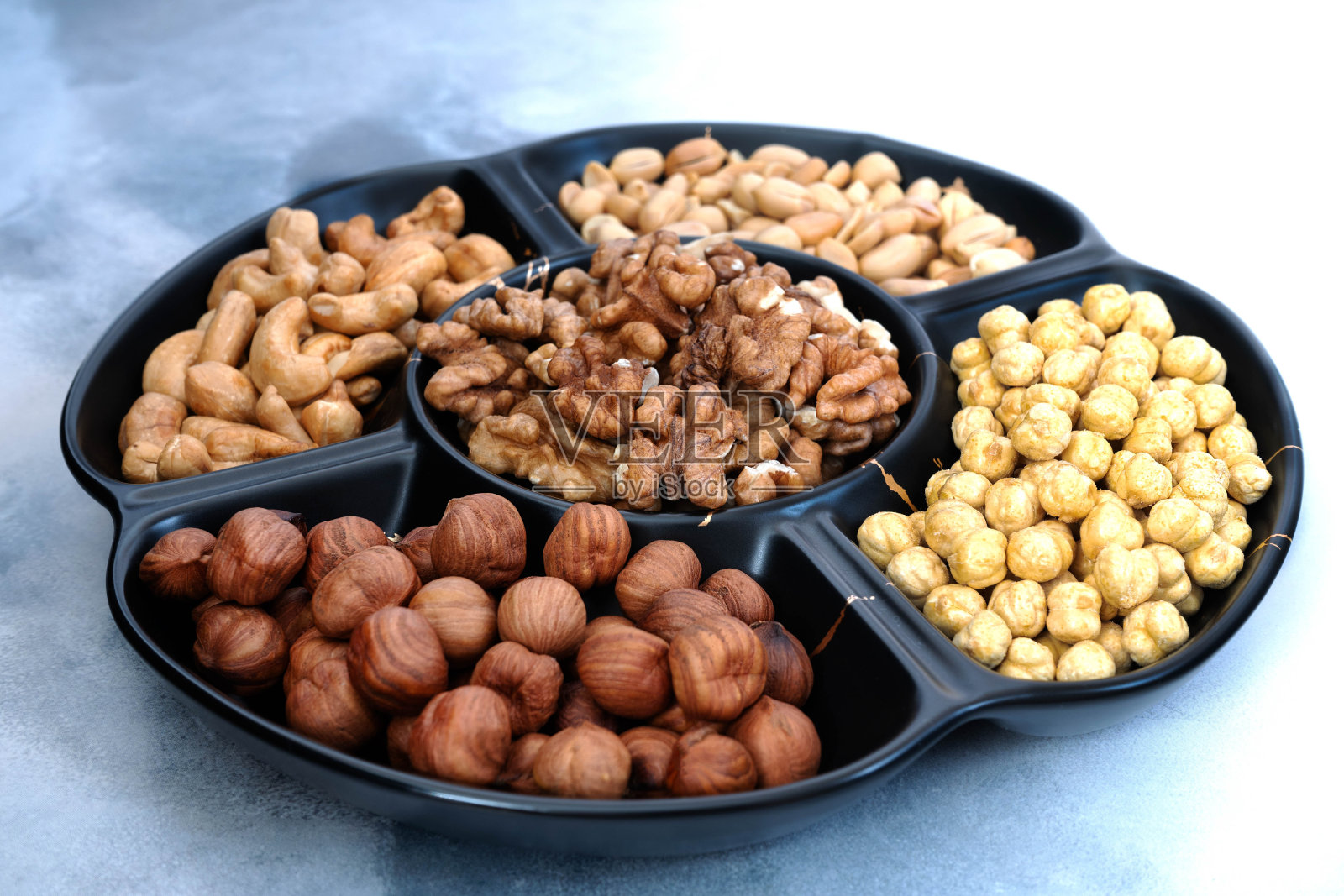 浅灰色的桌子上放着一个黑色的圆盘子，里面有五格各式各样的果仁(榛子、腰果、花生、核桃和鹰嘴豆)。特写镜头照片摄影图片