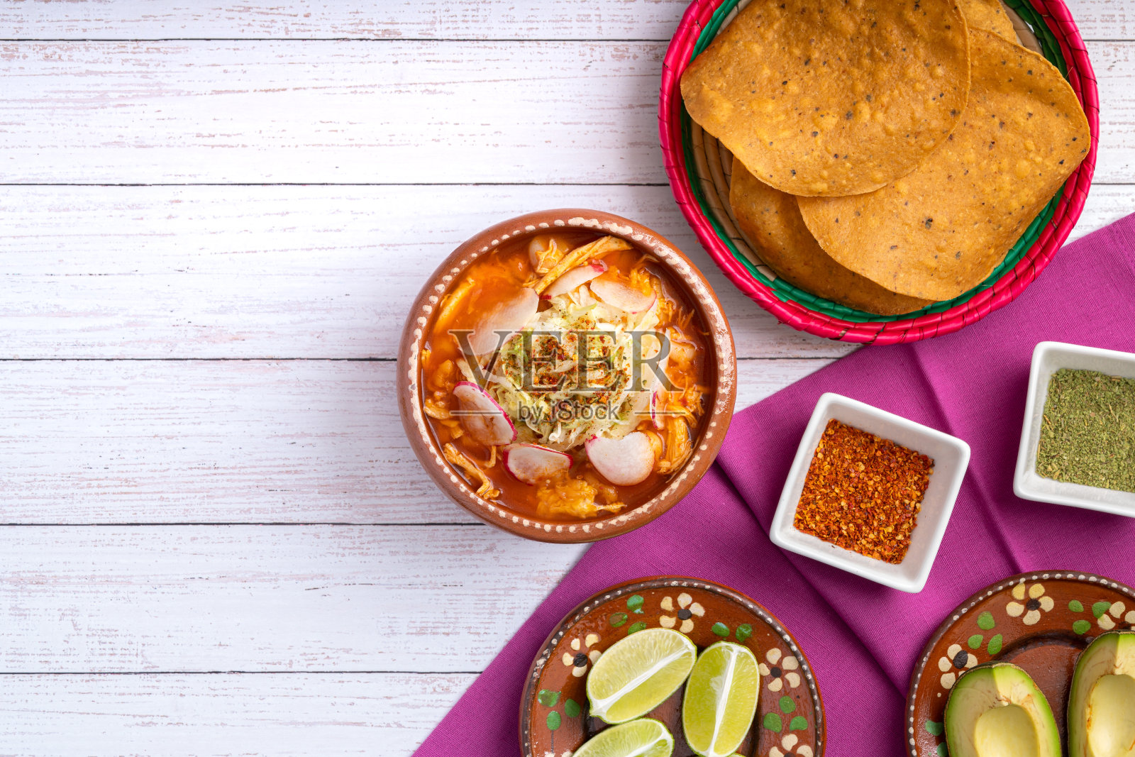 传统的墨西哥食物。红色的pozole鸡汤，配以牛油果、生菜、洋葱、萝卜、柠檬、辣椒和酥脆的玉米饼，也被称为tostadas，在白色木制的背景上。照片摄影图片