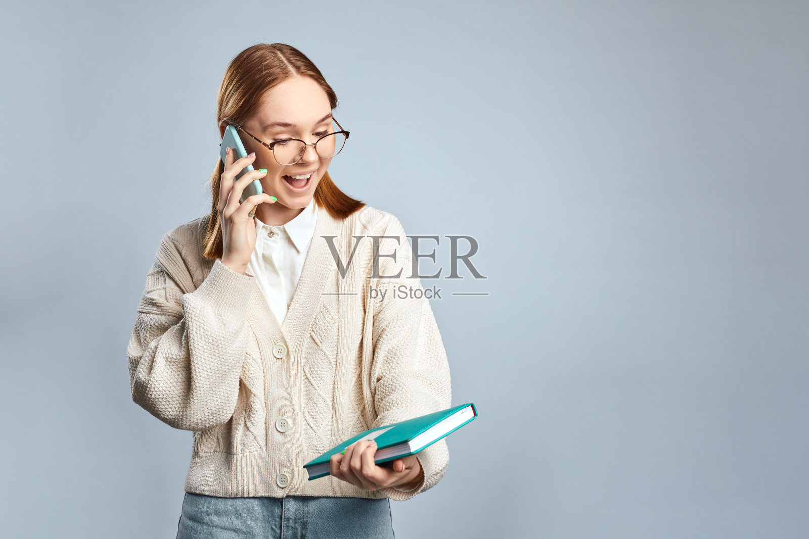 快乐的年轻女孩穿着休闲毛衣，一边读笔记本一边打手机。可爱的女学生用手机，手里拿着书。影棚拍摄。照片摄影图片