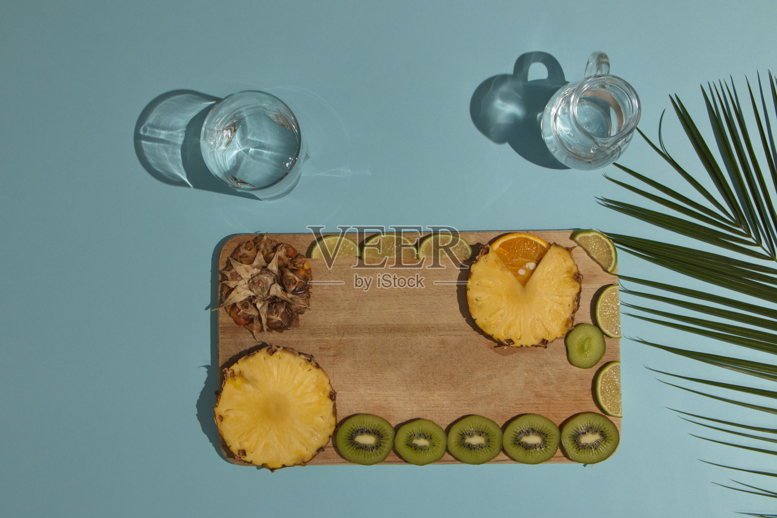 异国情调的夏季水果片木盘浅蓝色背景与棕榈叶和浅色调的眼镜。简约的平面场景。照片摄影图片