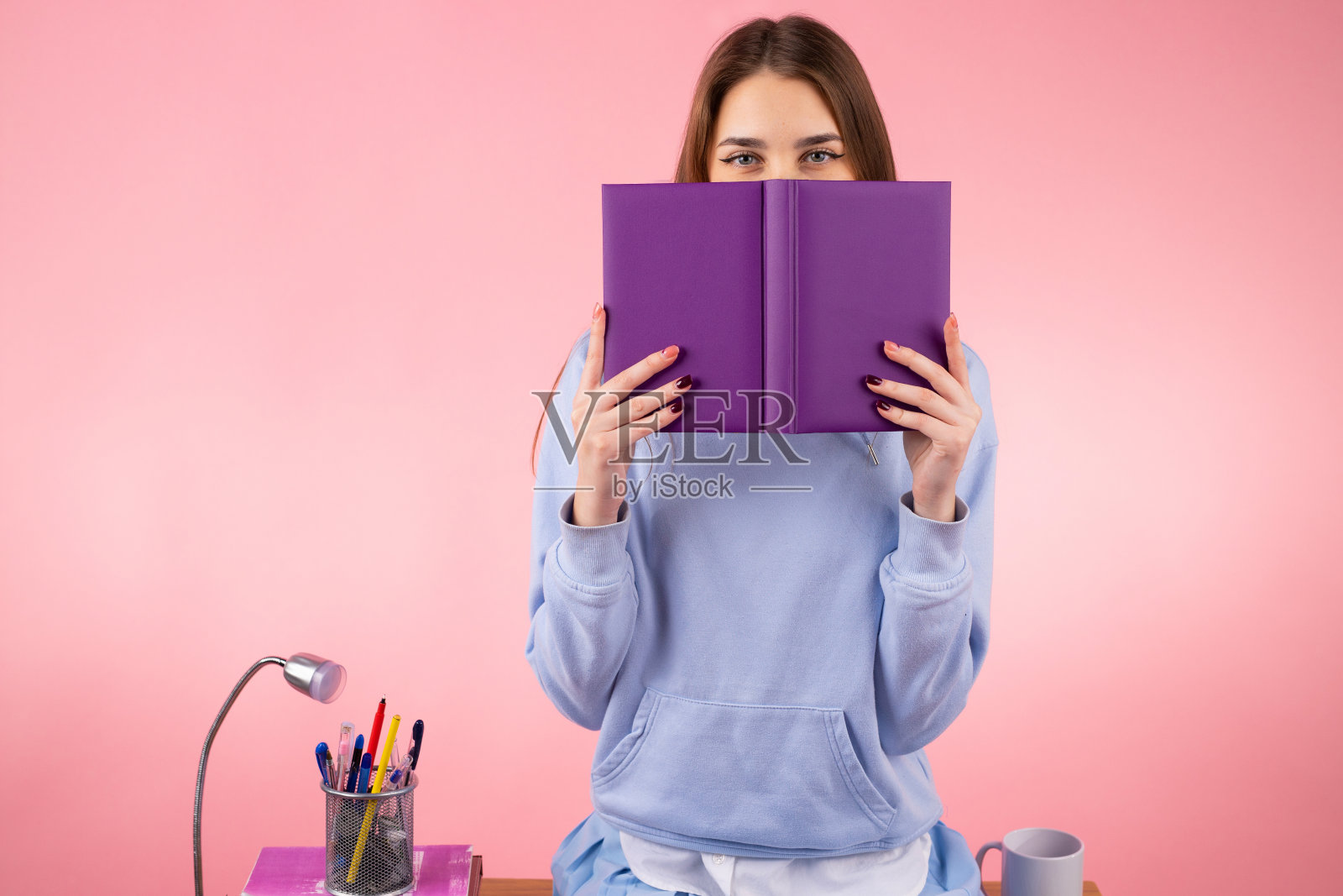 一个可爱的金发女孩坐在桌子上，用一本紫色的书盖住她的脸。照片摄影图片