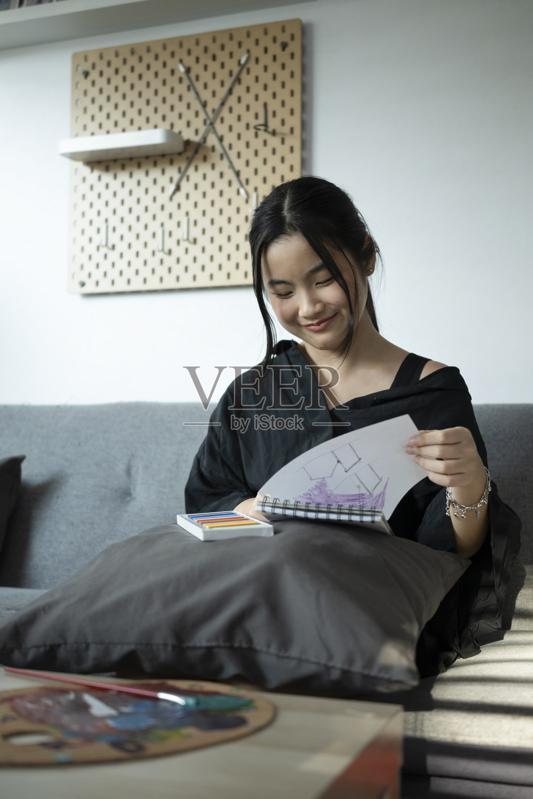 快乐的亚洲女孩画在她的笔记本，而坐在沙发上的客厅。照片摄影图片