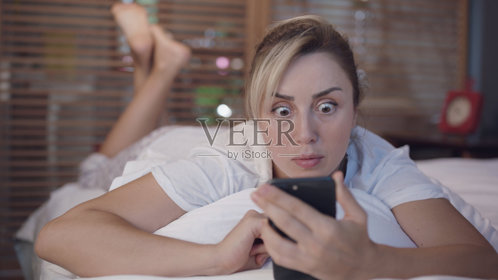 这名女子脸朝下躺在床上，盖着白色床单，她对手机上看到的东西感到惊讶。照片摄影图片