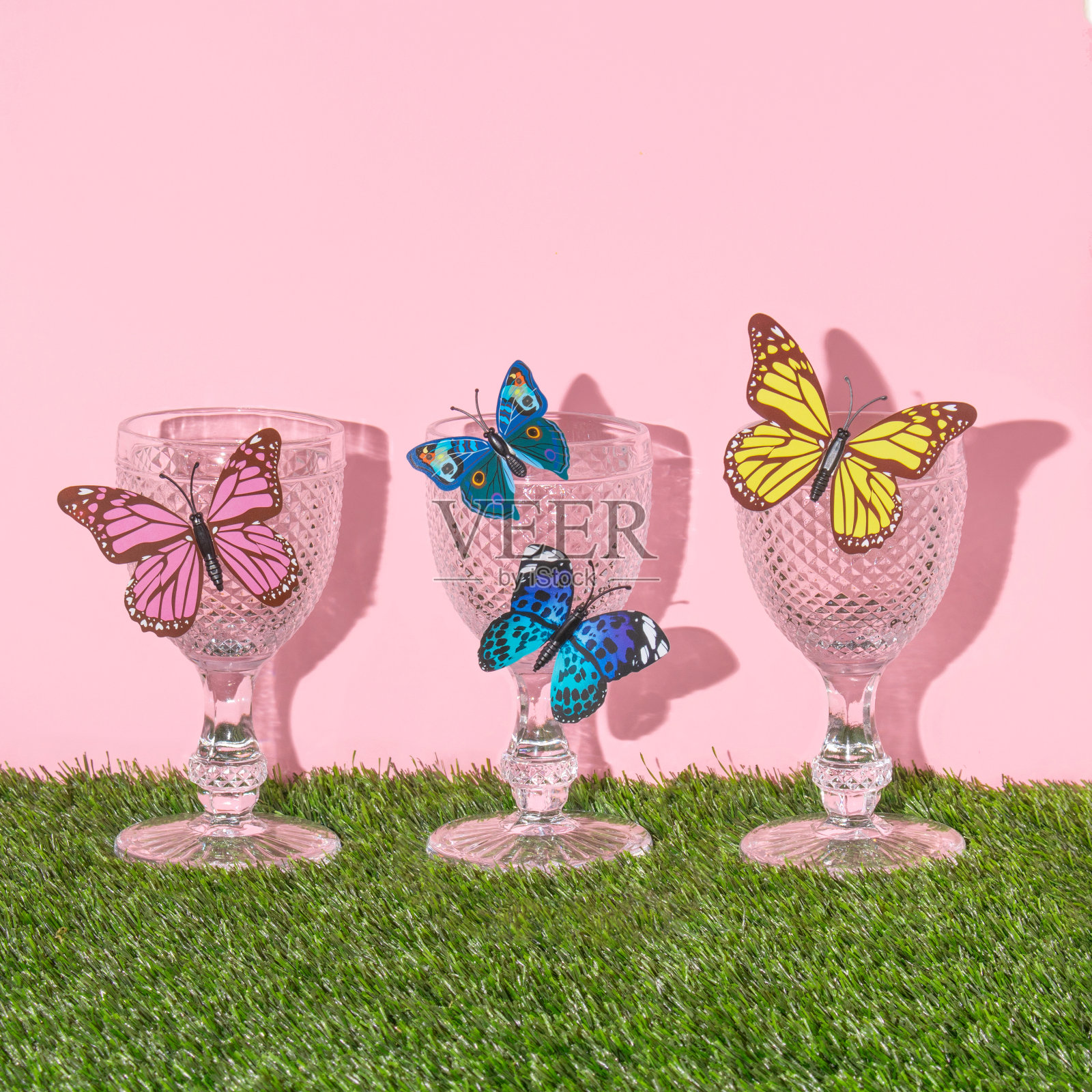 春天的创意布局，复古的酒杯和彩色的蝴蝶在柔和的粉红色背景和绿色的草地。80年代或90年代复古时尚美学夏日概念。最小浪漫的庆祝想法。照片摄影图片