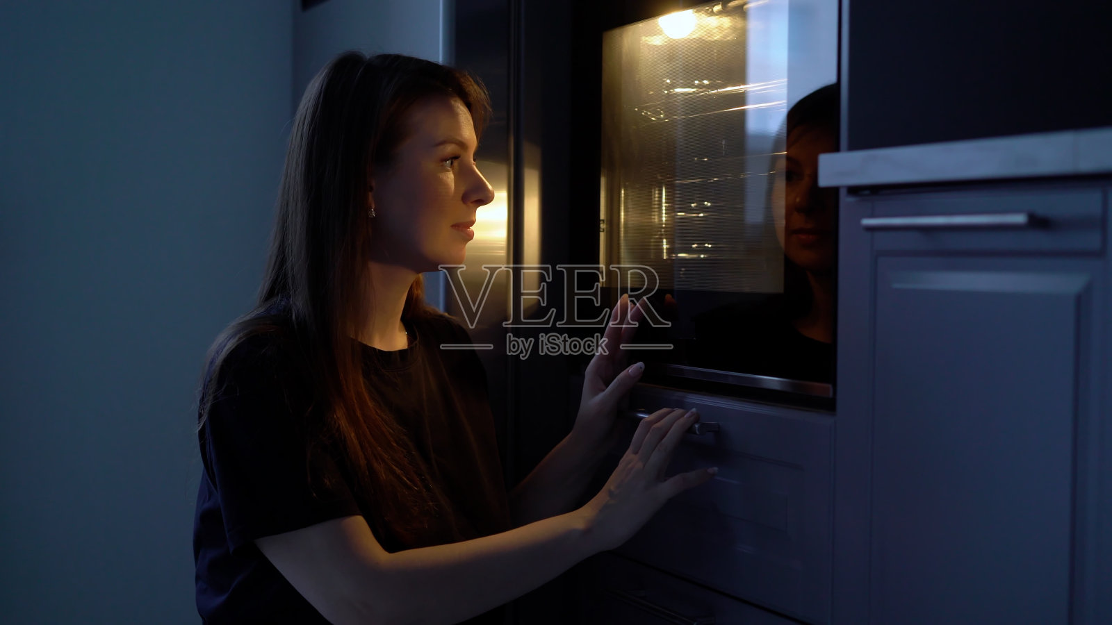 厨房里的一个年轻女人正在用烤箱做饭。烤箱发出的光照片摄影图片