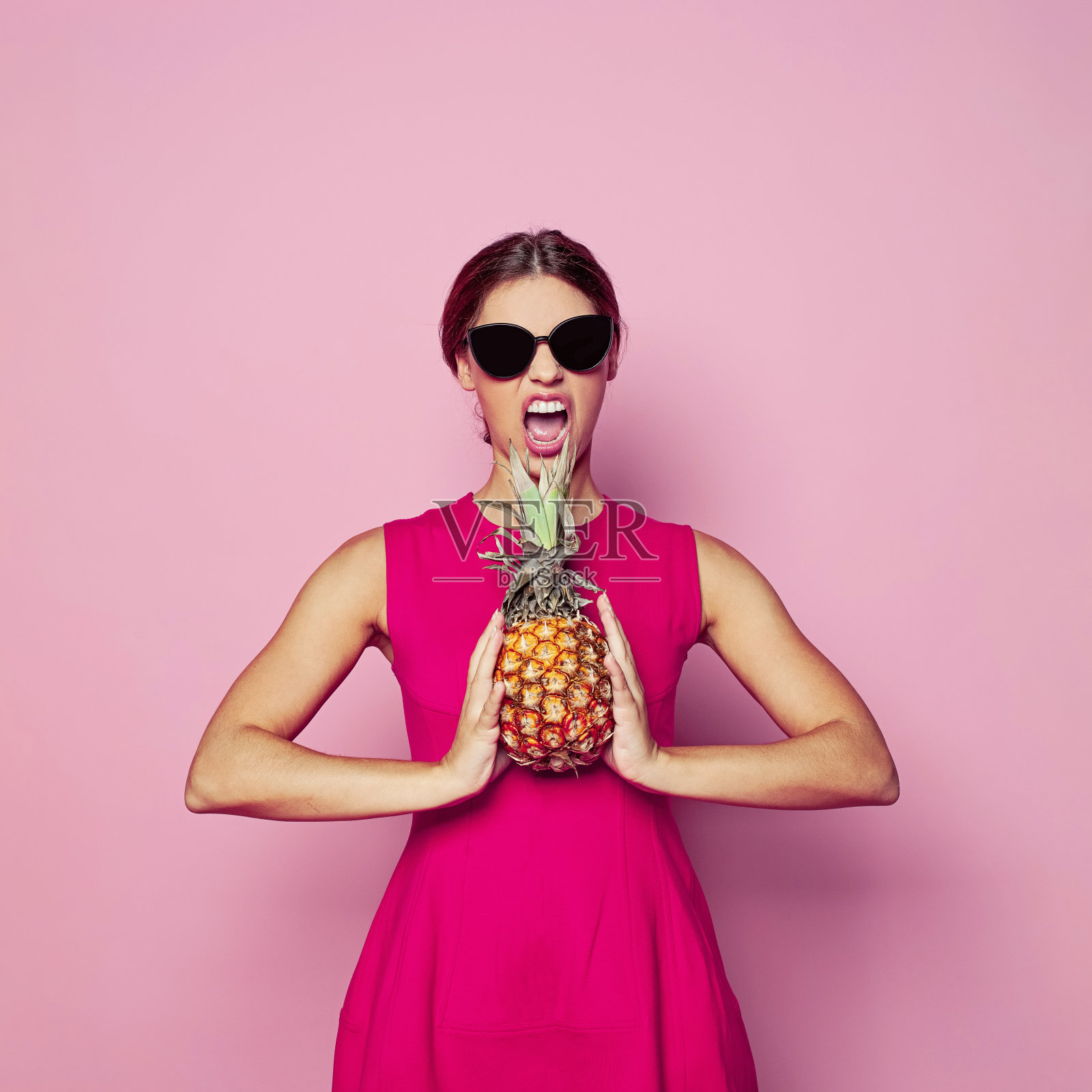 美丽的感性的女人在太阳镜拿着菠萝在粉红色背景的时尚肖像照片摄影图片