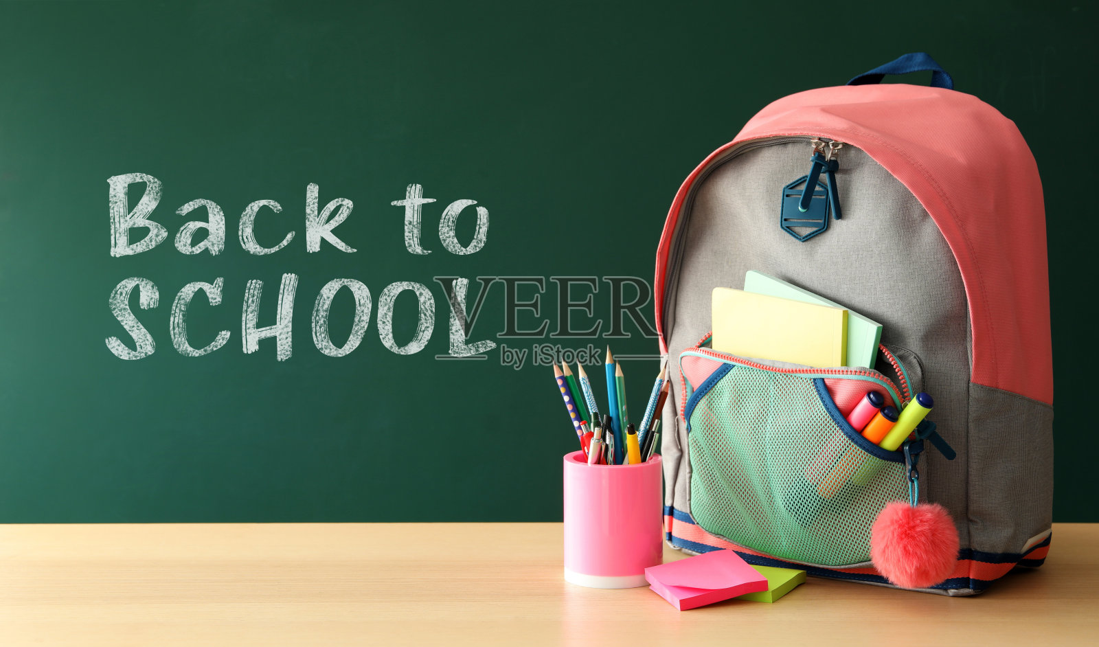 书包和不同的文具在木桌附近的绿色黑板上写着“回到学校”照片摄影图片