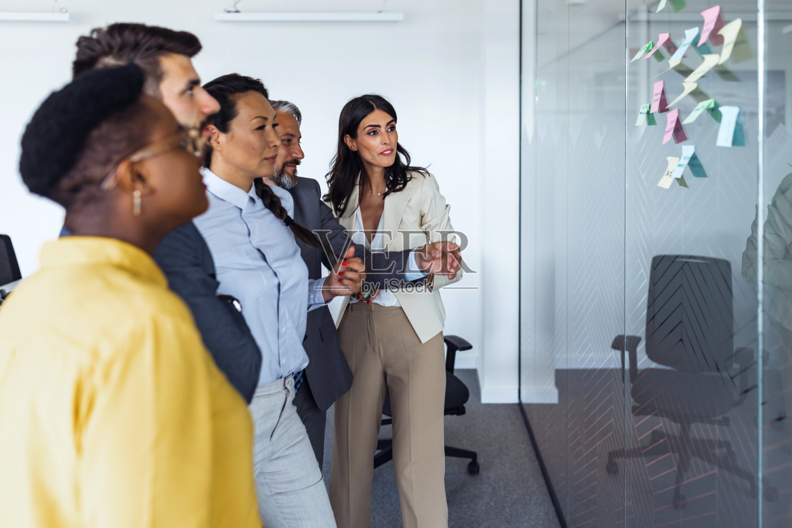 一群正在开会的商务人士站在明亮的办公室窗户前，看着用别针钉住的笔记照片摄影图片