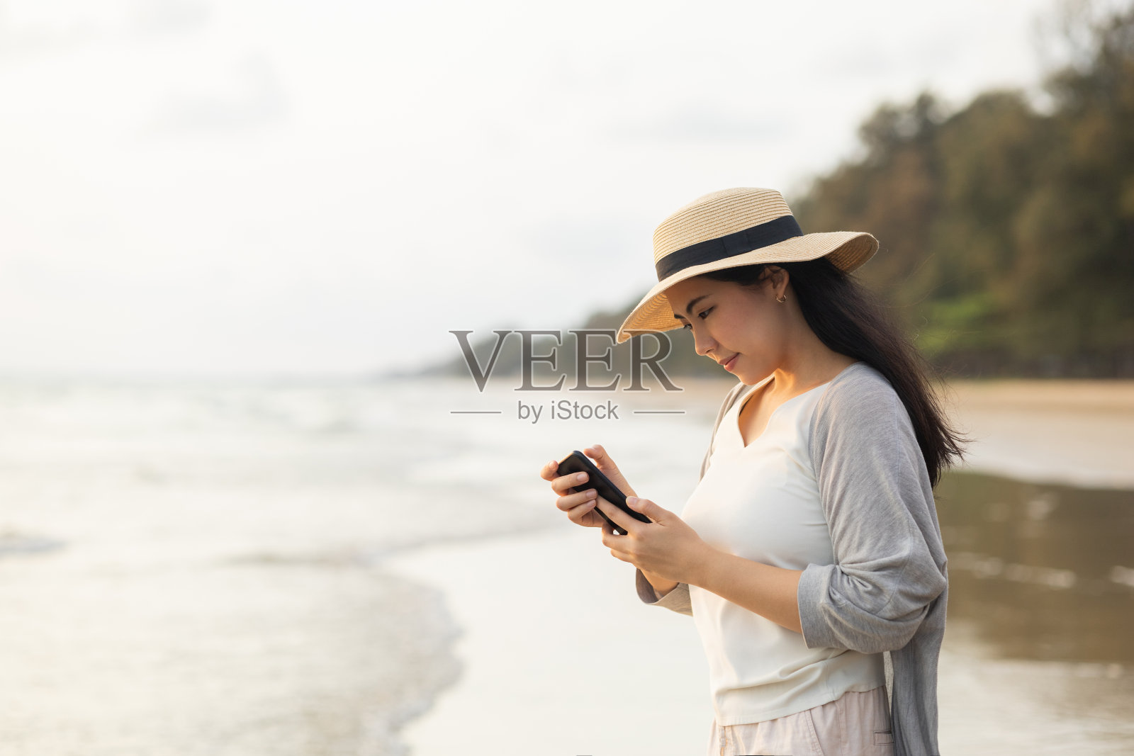 年轻的亚洲女性在度假期间在海滩上散步时使用智能手机。女性在海边旅行时用手机短信交流社交。照片摄影图片