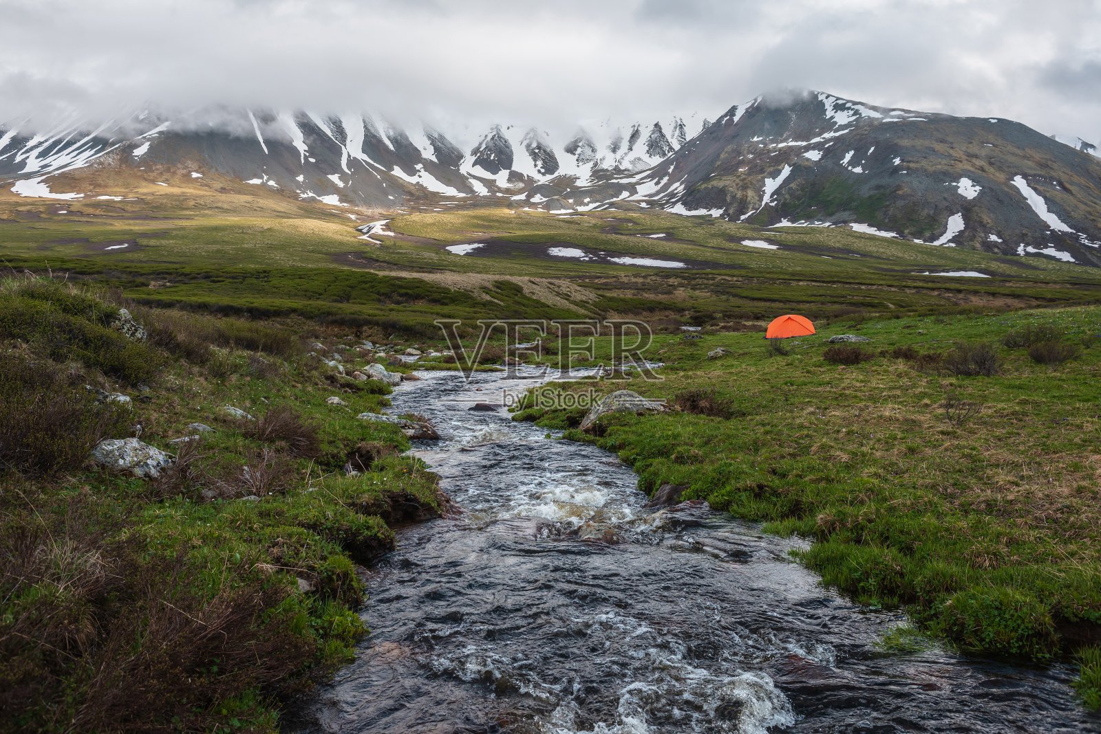 橙色帐篷，靠近清澈的小溪，可以看到低低的雨云中的山脉。引人注目的高山景观，山溪和雪山在灰色低云。苔原的阴森景色。照片摄影图片