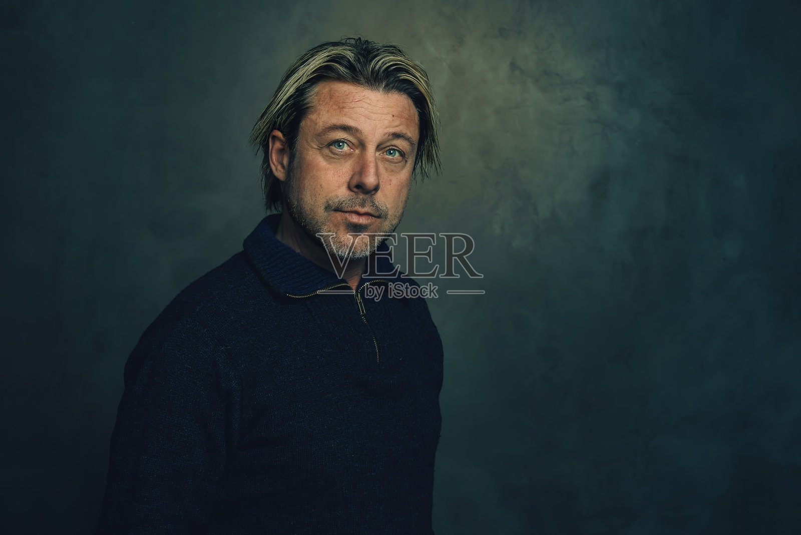 一个粗犷的留着胡茬的金发男人，穿着一件拉链领的蓝色羊毛衫，站在一堵深灰色的墙前。照片摄影图片