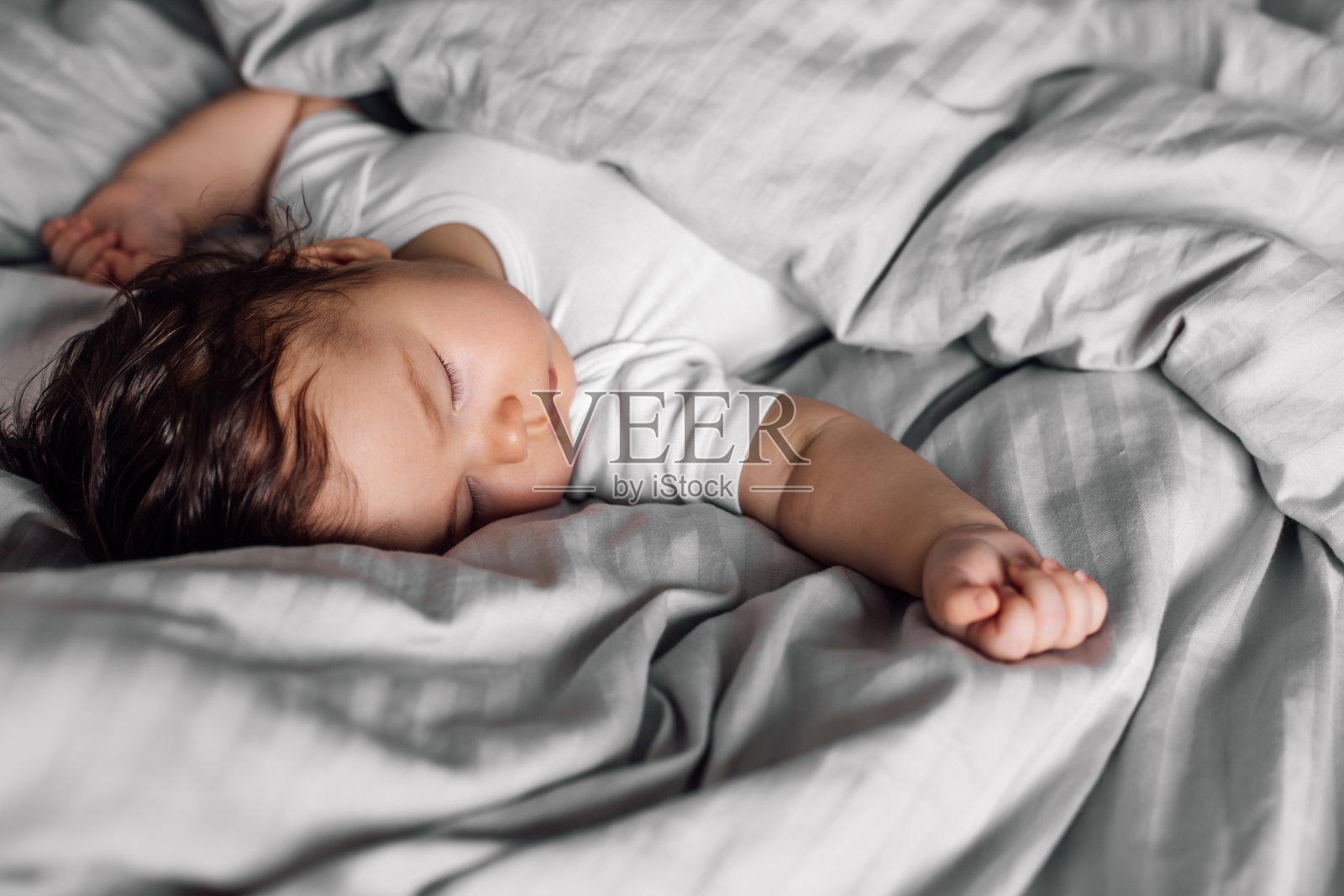 漂亮的婴儿穿着睡衣躺在家里的床上，盖着灰色的毯子。婴儿在卧室里熟睡，看到甜美的梦。灰色背景，自由复制空间。儿童保育与健康睡眠理念照片摄影图片