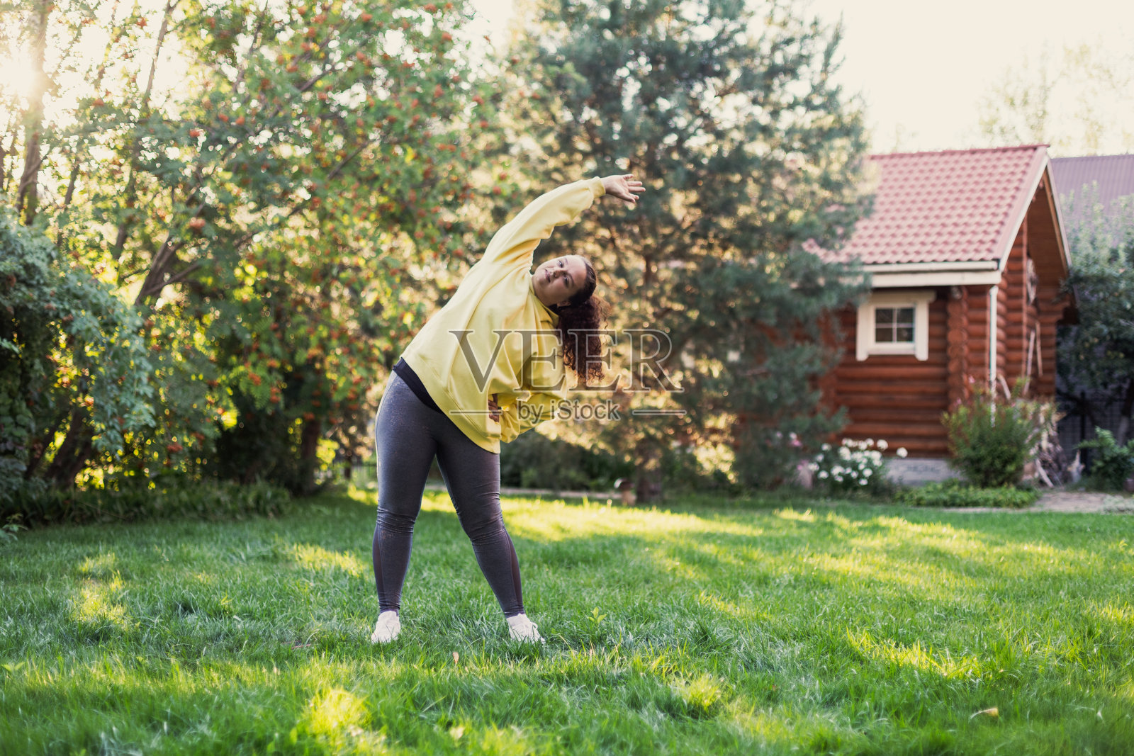 女人在绿色的草地上伸展手臂，穿着运动服，看着后院的摄像机，背景是乡村别墅和高大的树木。身体正照片摄影图片
