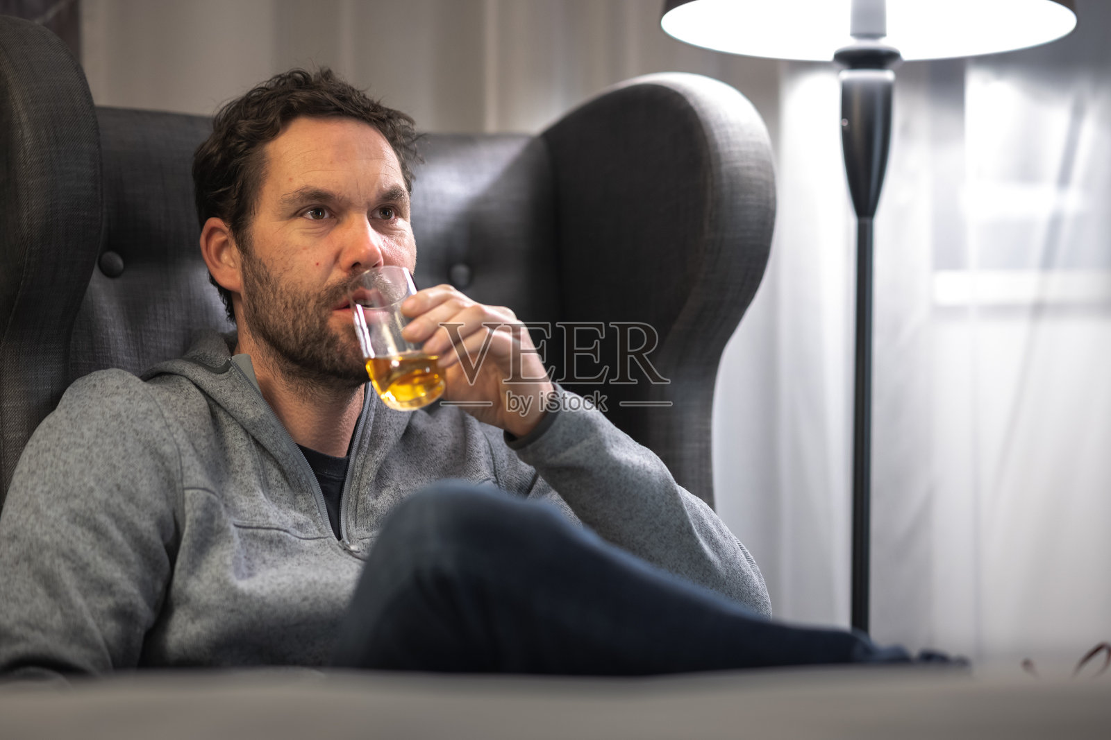 年轻的潮人面无表情，在酒店房间里用杯子喝着酒精饮料。照片摄影图片