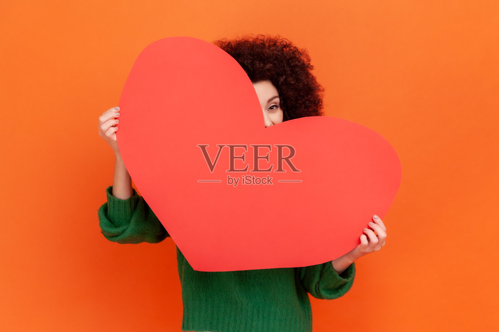 俏皮浪漫的女人，非洲式发型，穿着绿色休闲风格的毛衣，从大红心偷窥，调情，表达爱。照片摄影图片