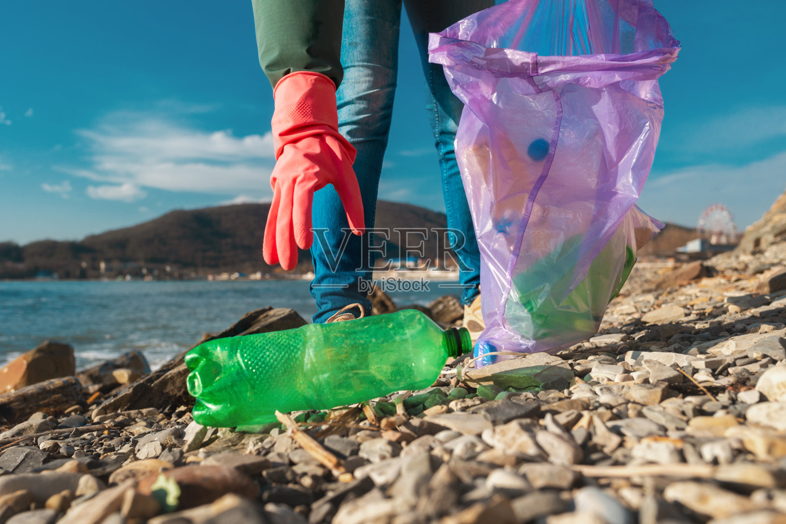 一位戴着橡胶手套的志愿者伸手去拿躺在海边的一个脏塑料瓶。手的特写镜头。环境污染照片摄影图片