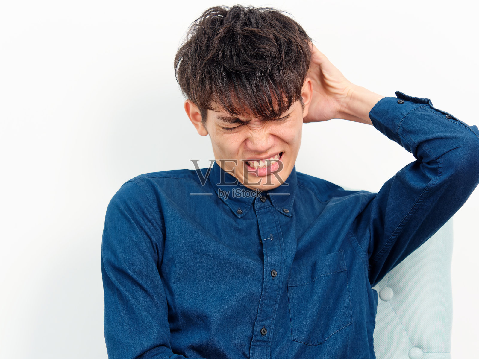 英俊的中国年轻男子的肖像与卷曲的黑色头发，蓝色衬衫坐在扶手椅和摆姿势的白墙背景。擦着他的头发，看起来很焦虑，摄影棚里的正面镜头。照片摄影图片