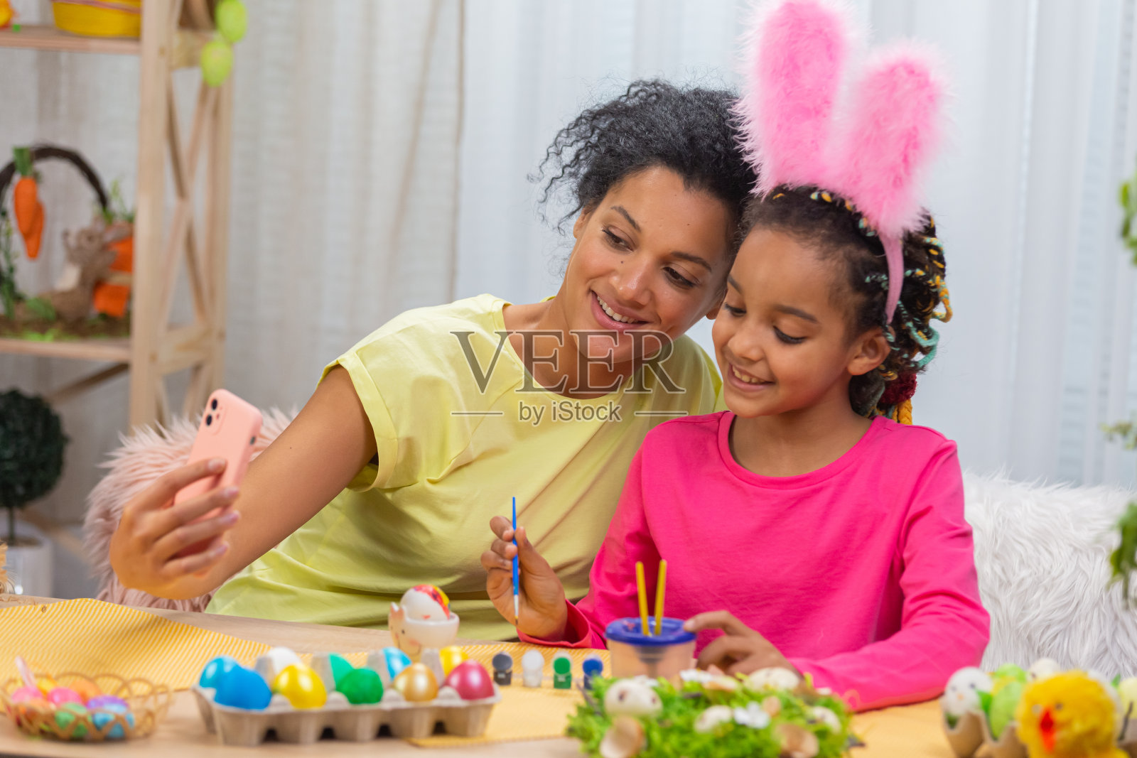 妈妈和长着滑稽兔子耳朵的女儿在用电话进行视频通话。非裔美国妇女和小女孩坐在节日装饰的房间里的桌子旁。复活节快乐。近距离照片摄影图片