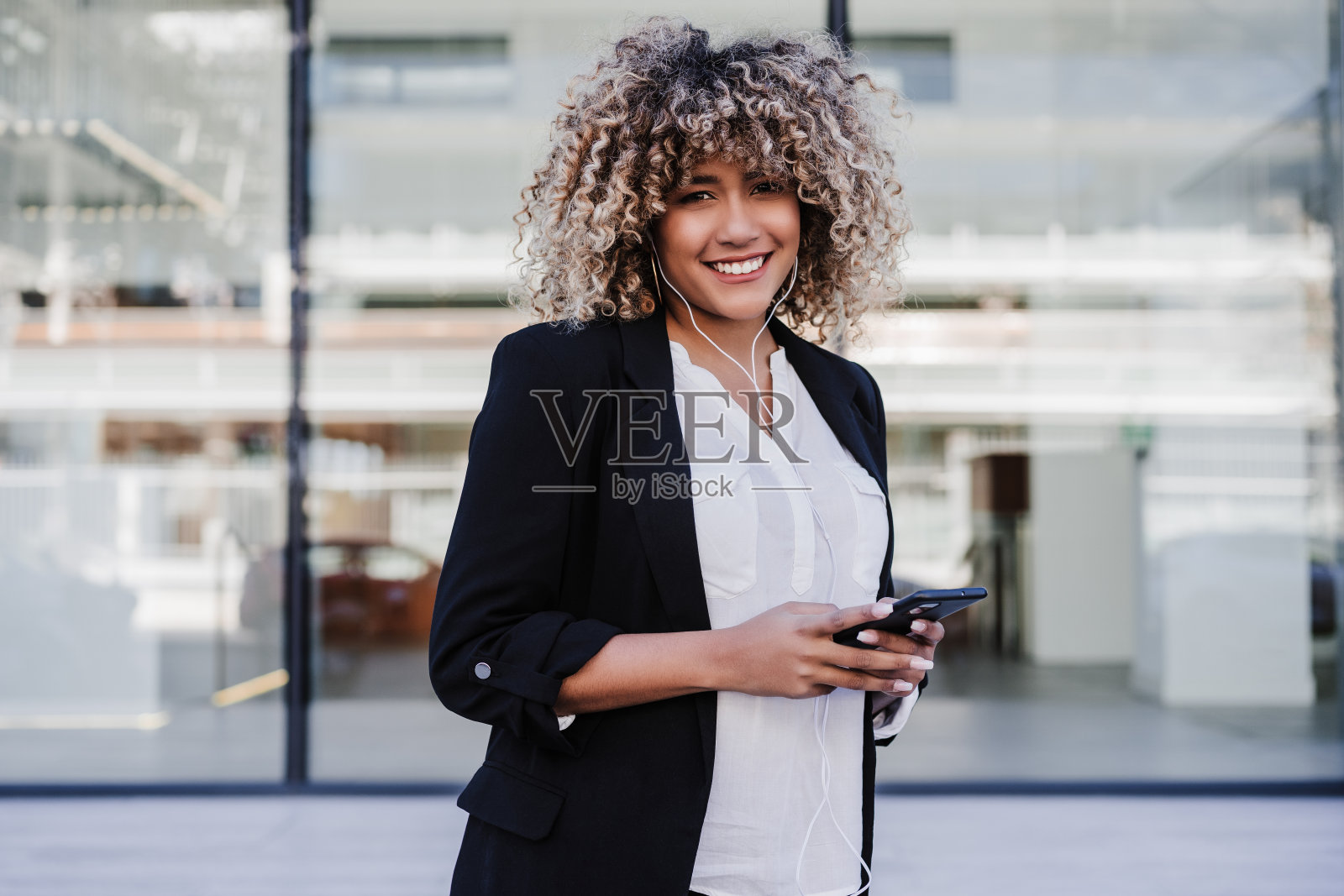 美丽的微笑商业妇女使用手机和耳机在城市。建筑的背景照片摄影图片