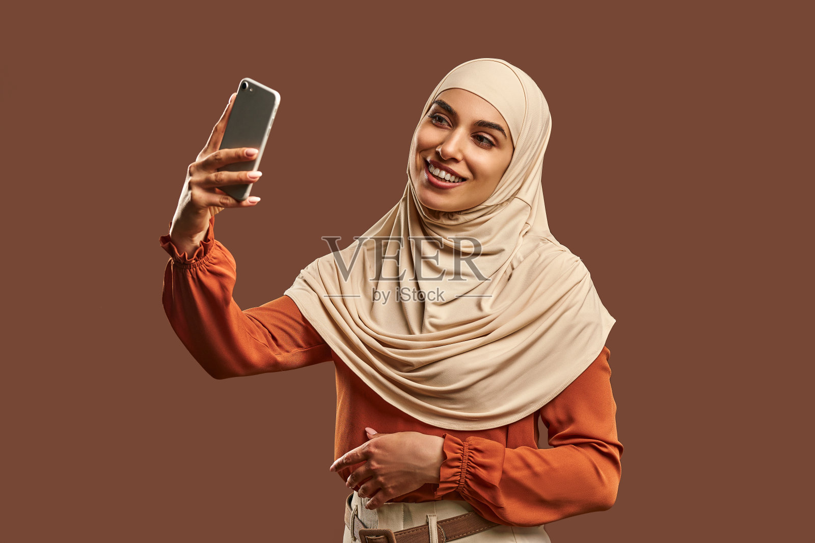美丽的穆斯林妇女，戴着米色头巾，手里拿着手机，微笑着站在棕色背景附近自拍。照片摄影图片