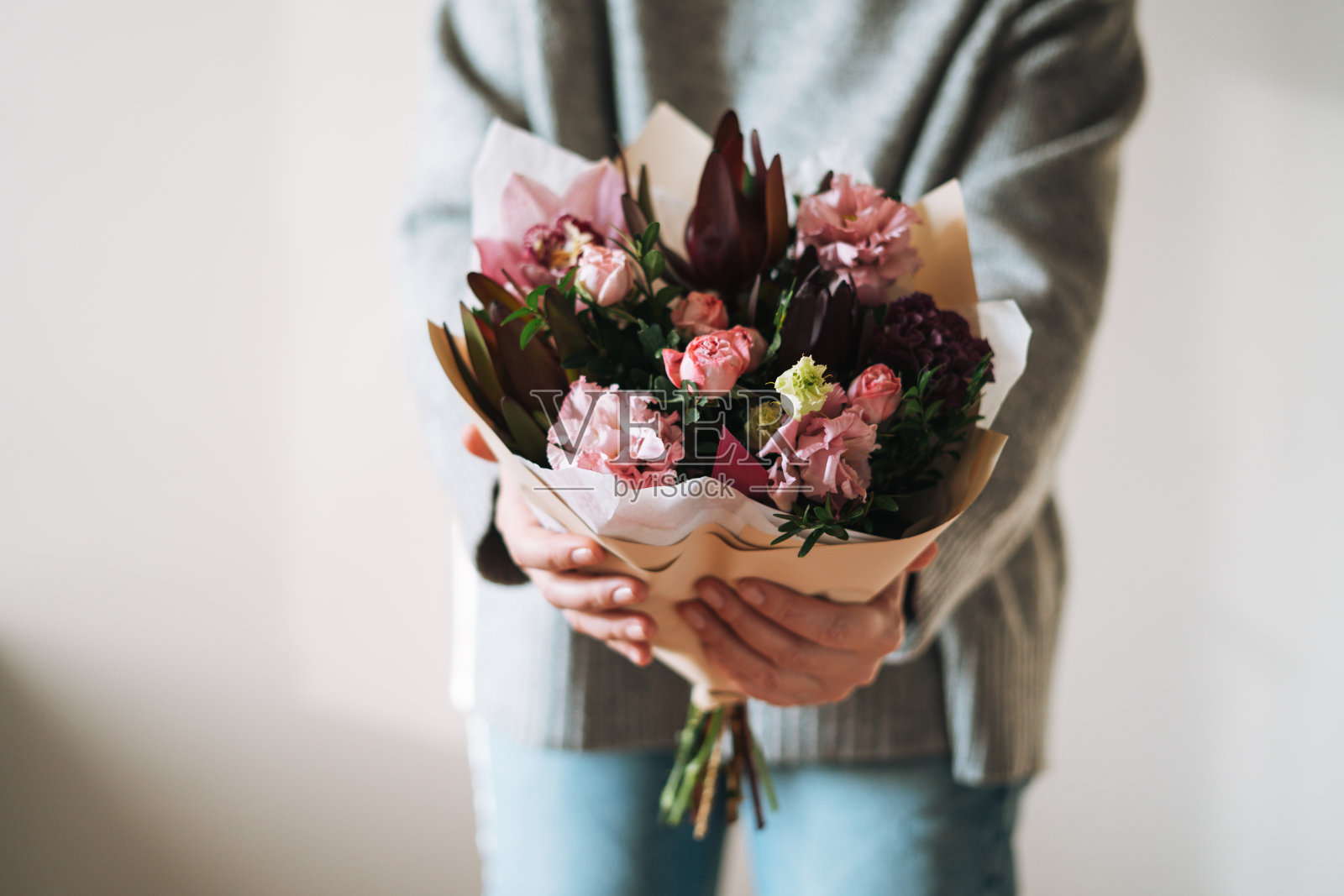 年轻女子身穿灰色针织毛衣，手里捧着一束不同的粉红色花朵，在灰色的背景上，选择焦点照片摄影图片