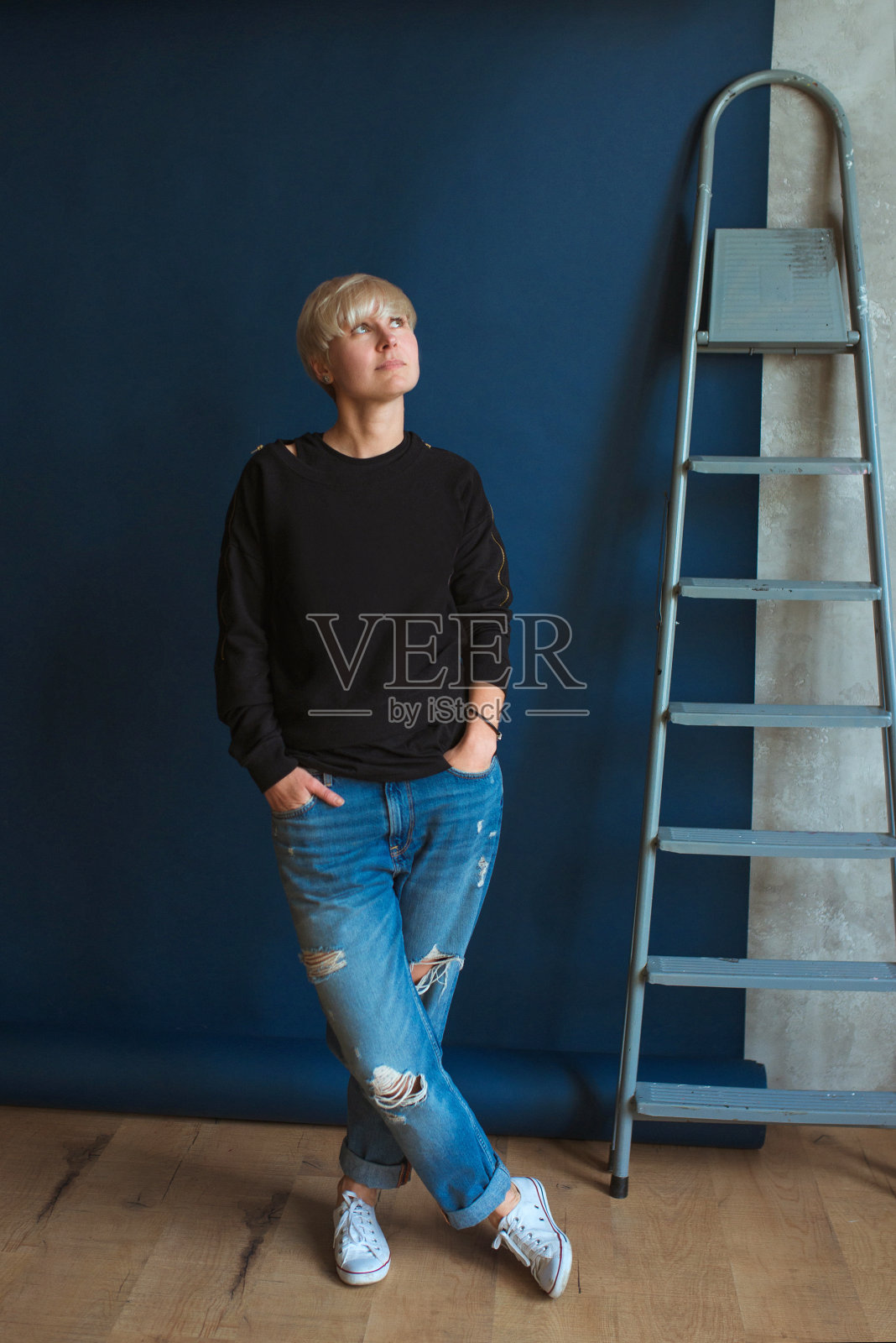 年轻的金发现代女性，穿着黑色运动衫和牛仔裤，站在梯子(楼梯)附近，计划翻修(修理)照片摄影图片