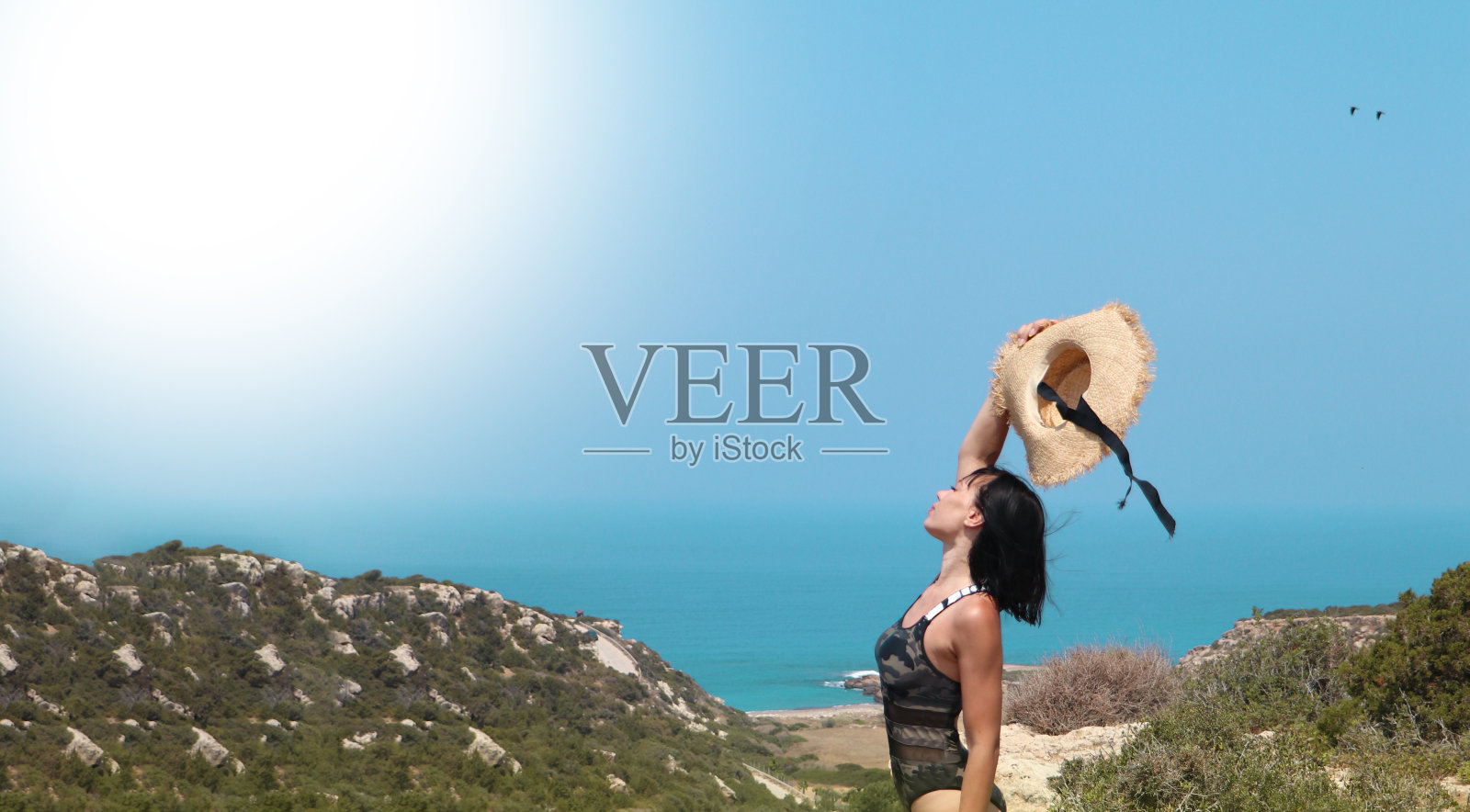 在大海和群山的映衬下，一位年轻女子举起了她的沙滩帽照片摄影图片