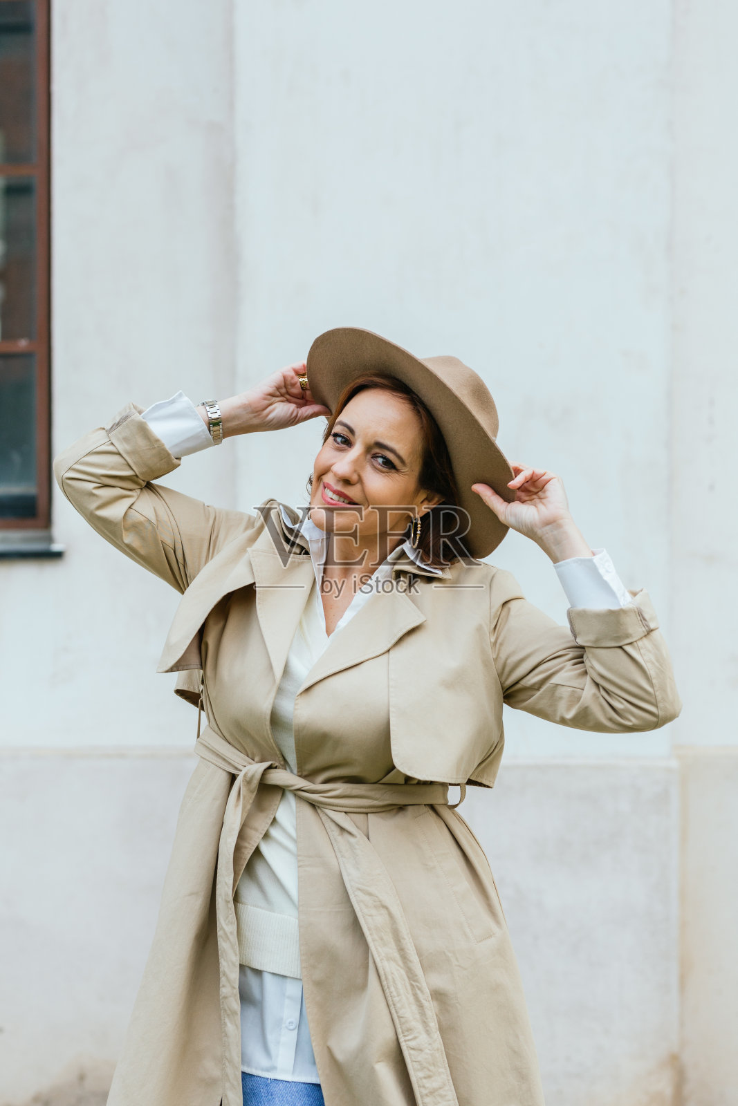 女人穿着时尚的风衣和帽子看着相机，而摆姿势在户外的街道上。照片摄影图片