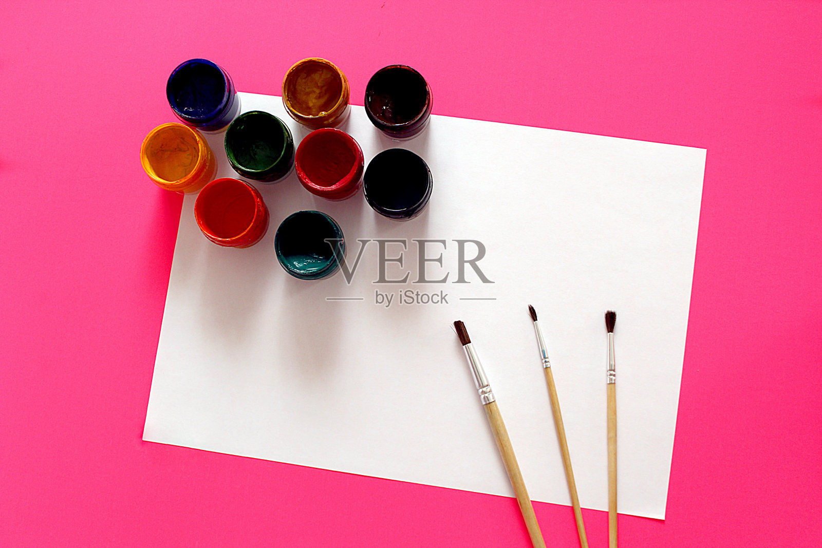 水粉画颜料和三种不同的笔刷与白纸躺在粉红色的背景上。照片摄影图片