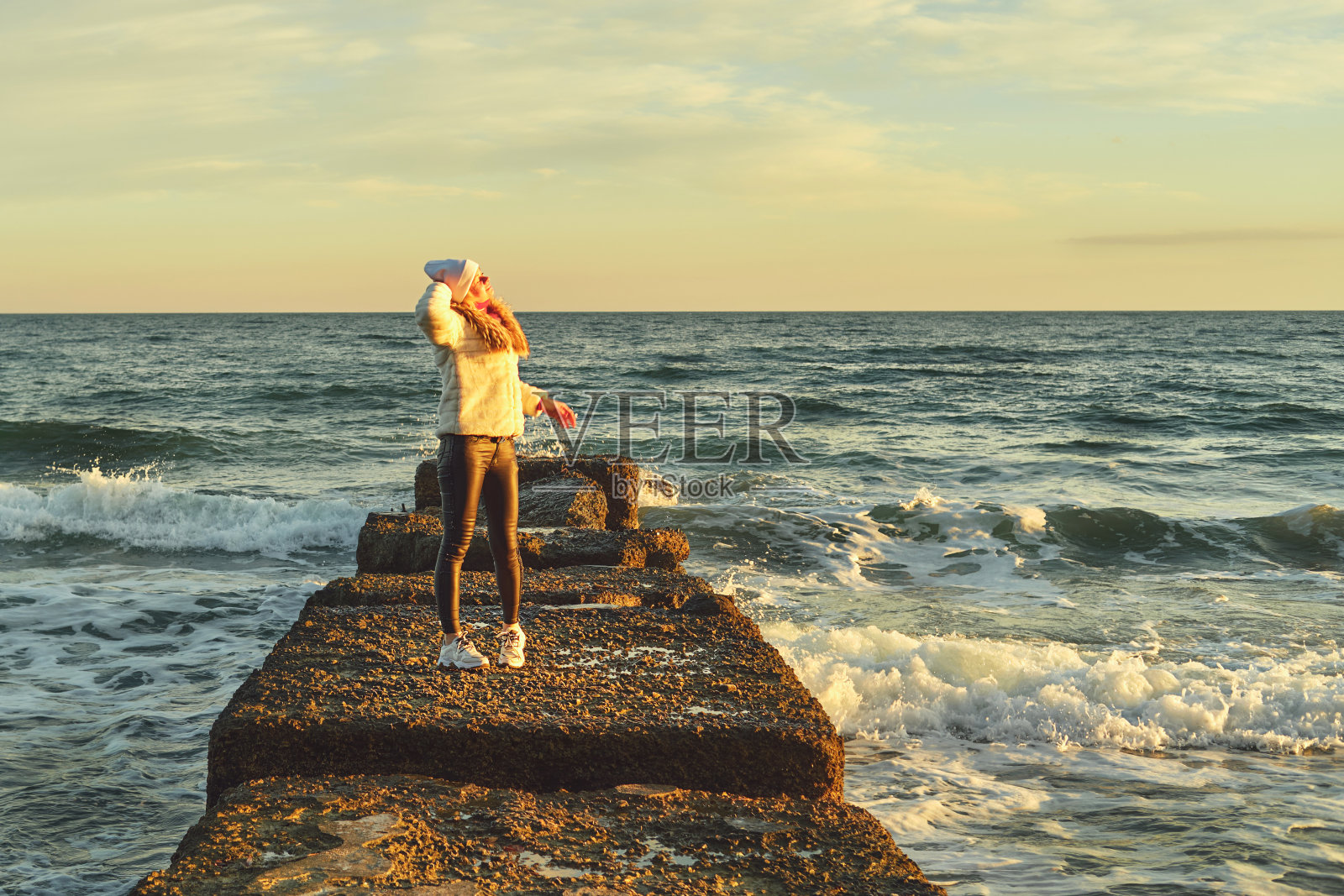 一个金发女人戴着帽子，穿着毛皮大衣，沿着海边的石堤走着照片摄影图片