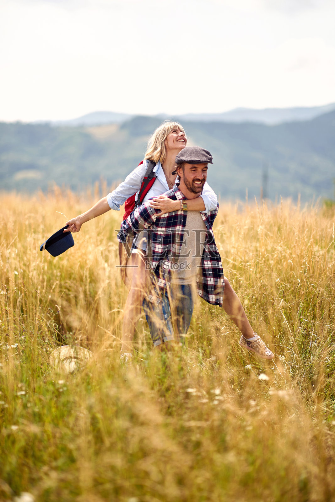 年轻快乐的高加索徒步旅行夫妇在田野微笑，一起有乐趣。女孩肩扛。运动、爱情、自由、假日的概念。照片摄影图片
