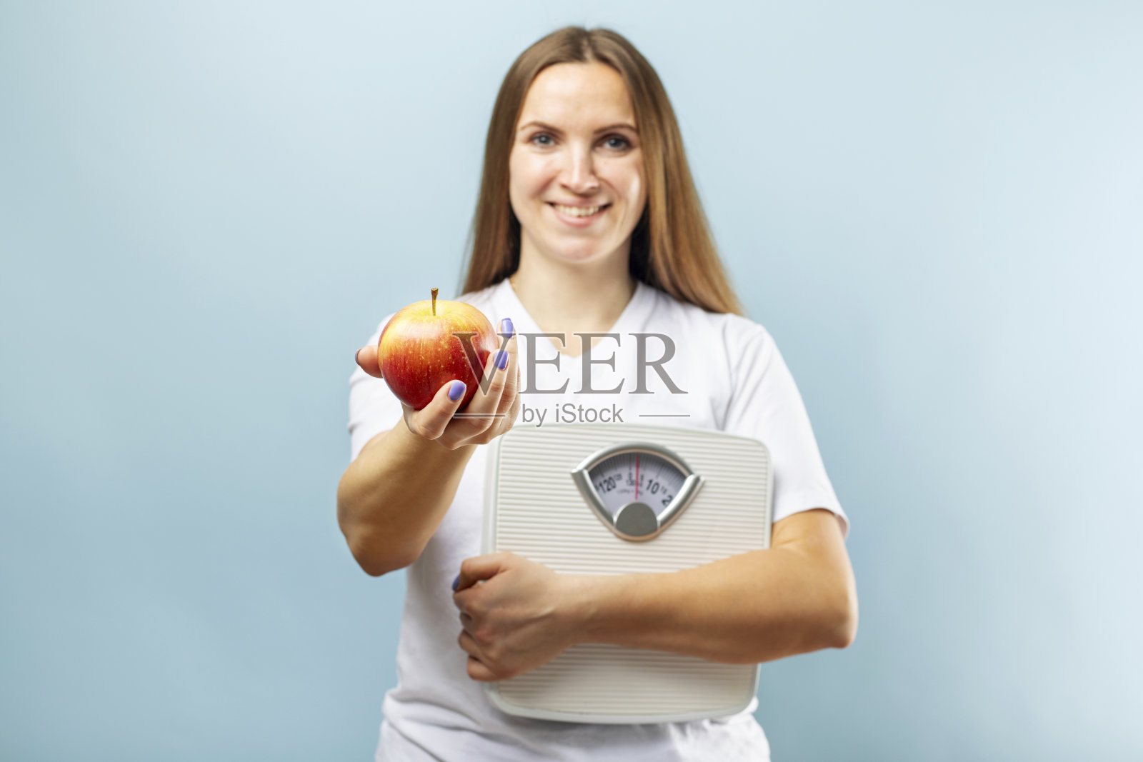 幸福微笑的女人拿着体重秤，蓝色背景上放着红苹果。健康饮食的概念。饮食照片摄影图片